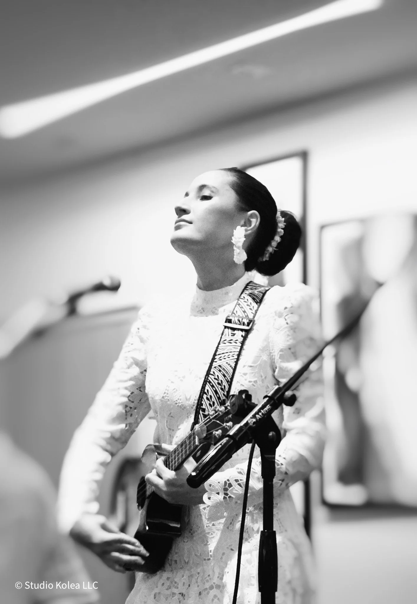 A woman with dark hair in a bun, wearing a white lace dress and large earrings, is playing an acoustic guitar during a performance.