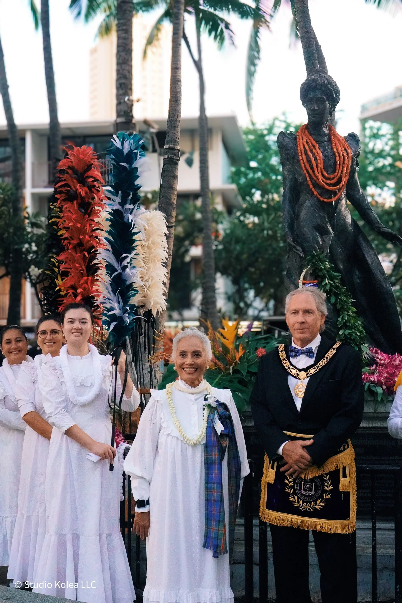 A parade float featuring a dark-colored statue of a woman adorned with orange necklaces. The float is decorated with colorful feathers and flowers. Several smiling women in white dresses stand in front of the float, with a woman in traditional attire