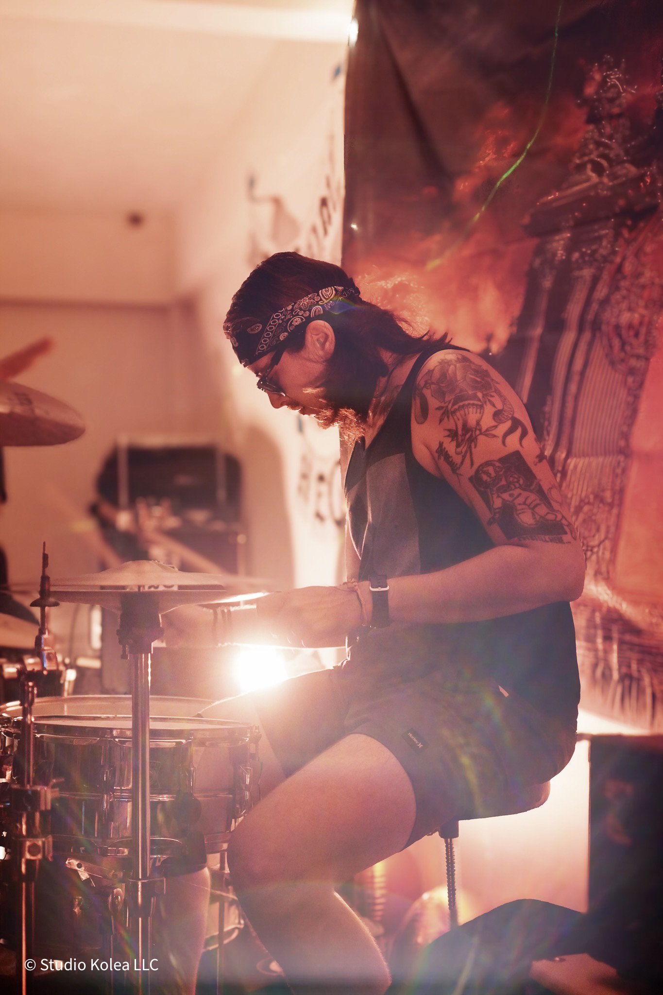 A person with a bandana and tattoos playing drums in a dimly lit room with bright backlighting.