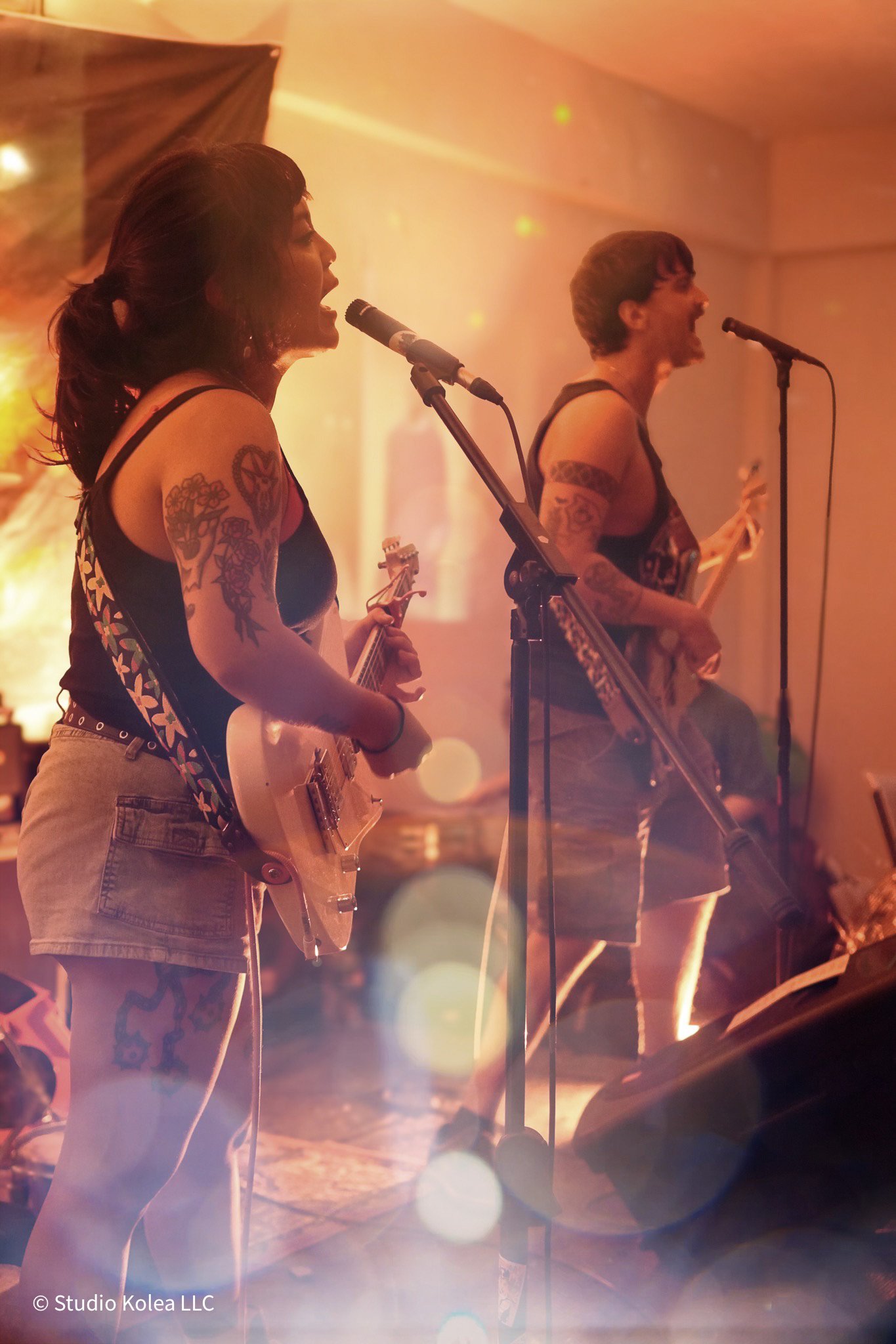 Two women with tattoos singing and playing electric guitars on stage during a live music performance, illuminated by warm stage lighting.