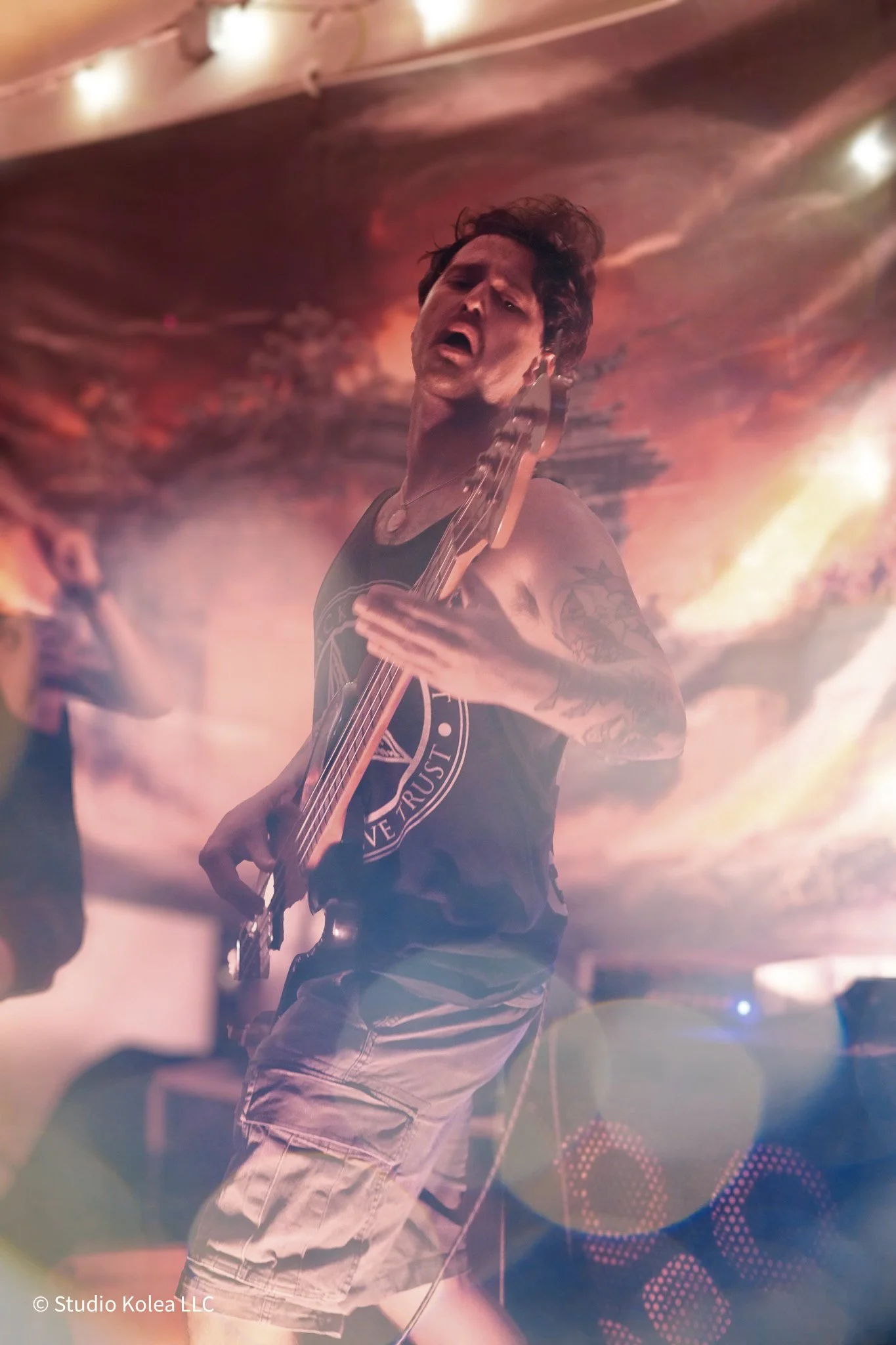 A male musician passionately plays an electric guitar on stage during a concert, with colorful stage lighting and a painted backdrop behind him.