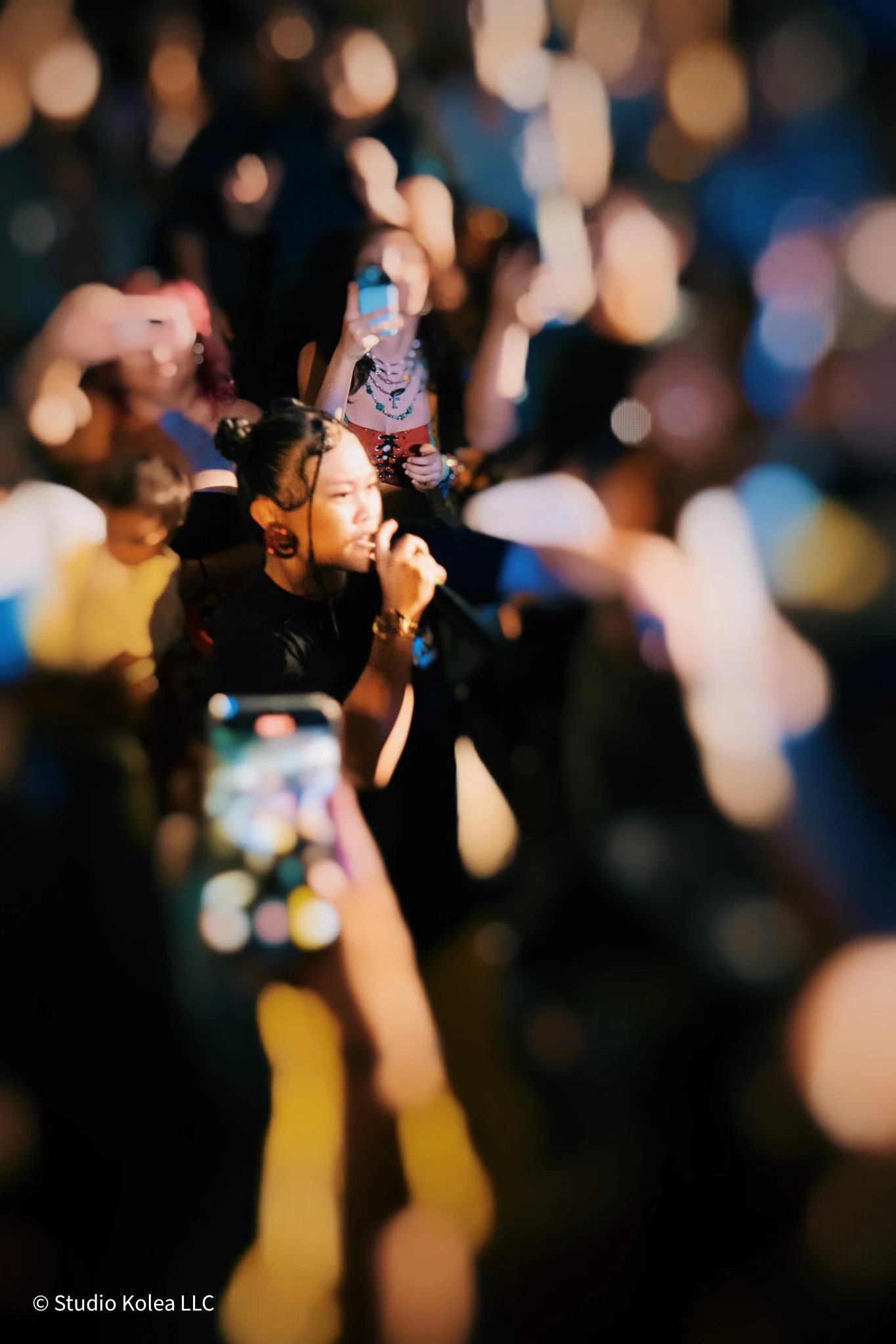 Crowd of people at an event, many holding up phones, with some taking photos or videos. The scene is illuminated with colorful, blurry lights in the background.