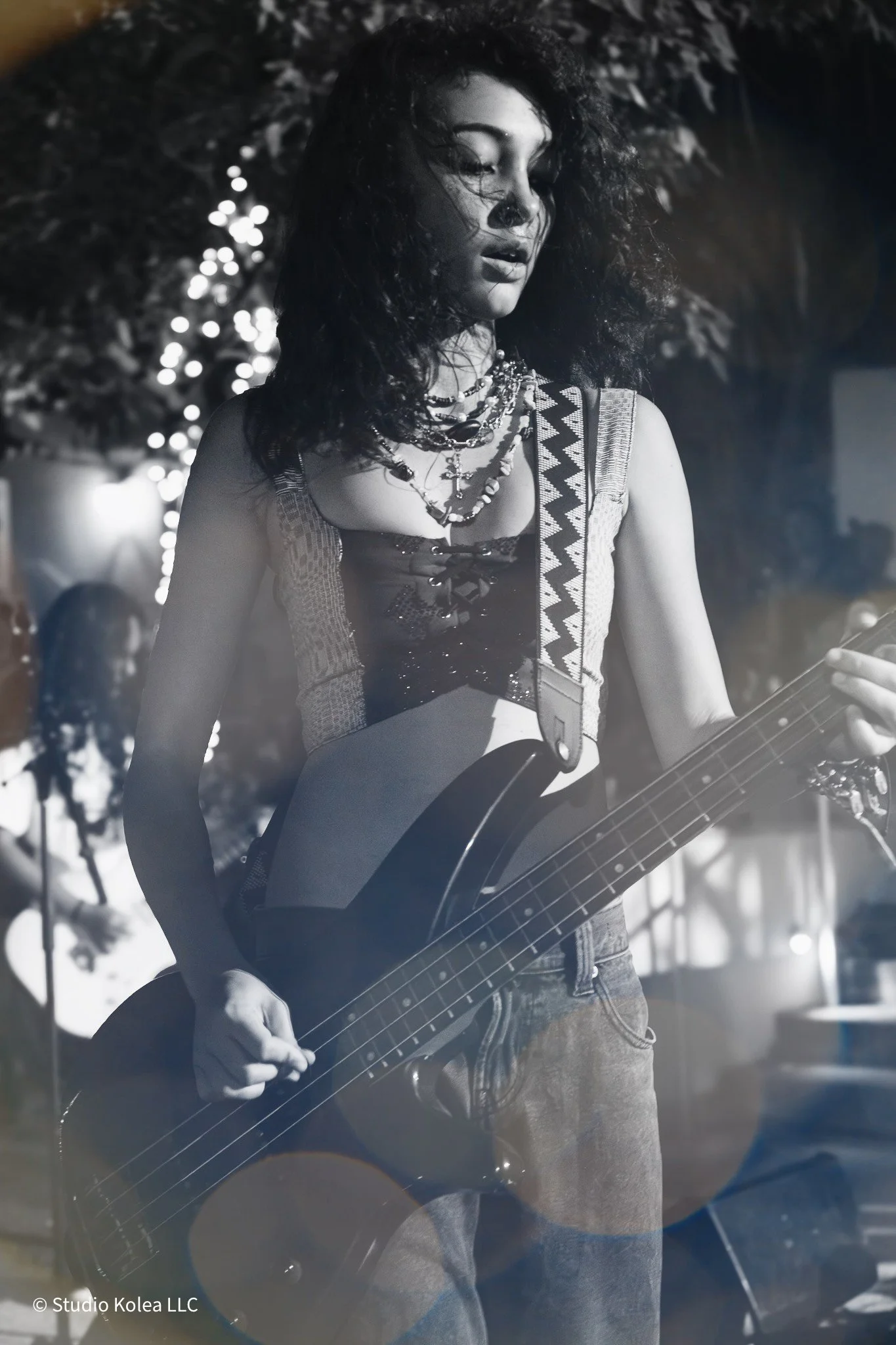 A woman with curly hair playing an electric guitar at a nighttime outdoor event, with trees and string lights in the background.