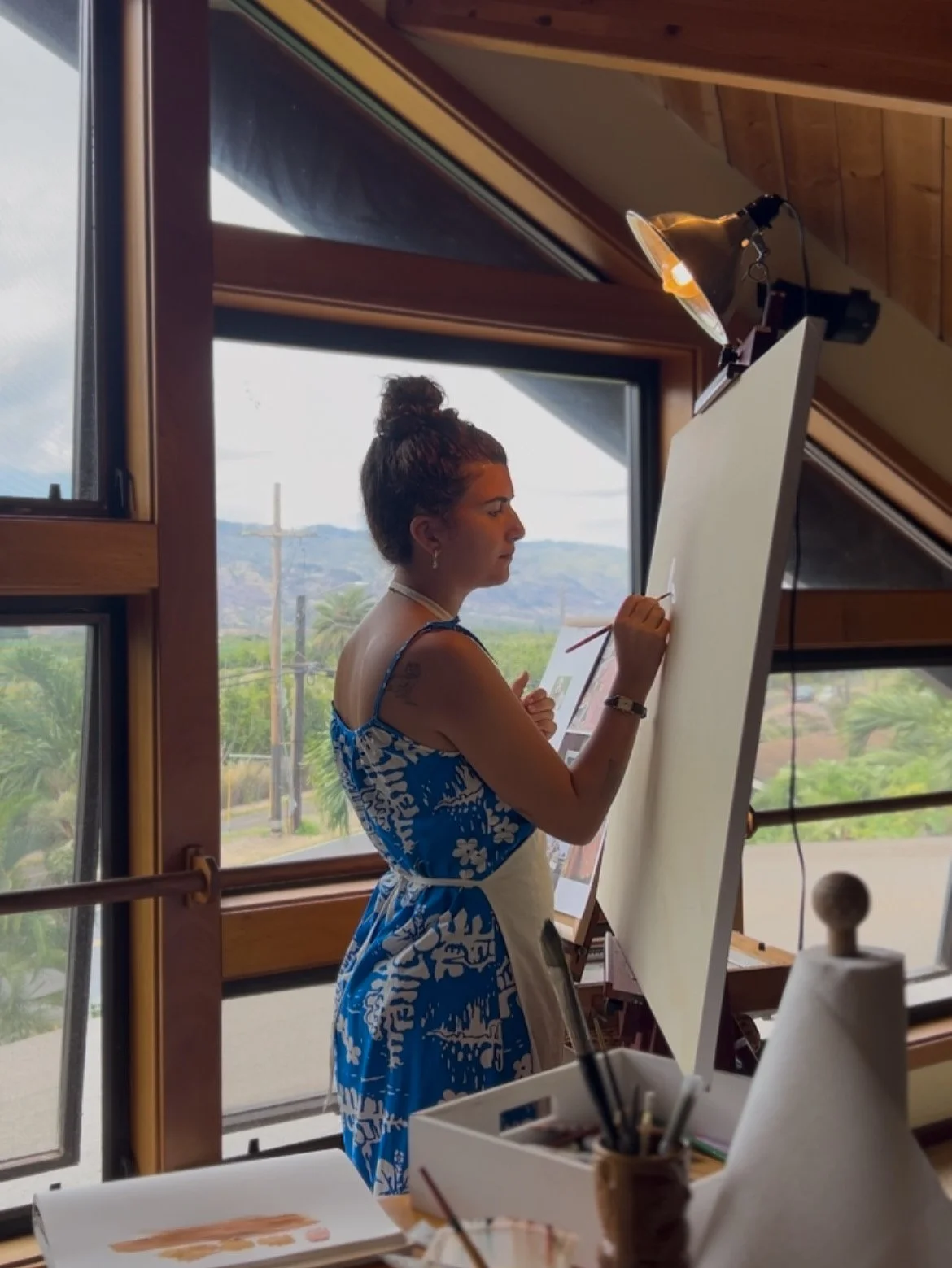 A woman in a blue and white floral dress stands at an easel, painting or drawing, in a room with large windows and wooden accents, overlooking a landscape outside.