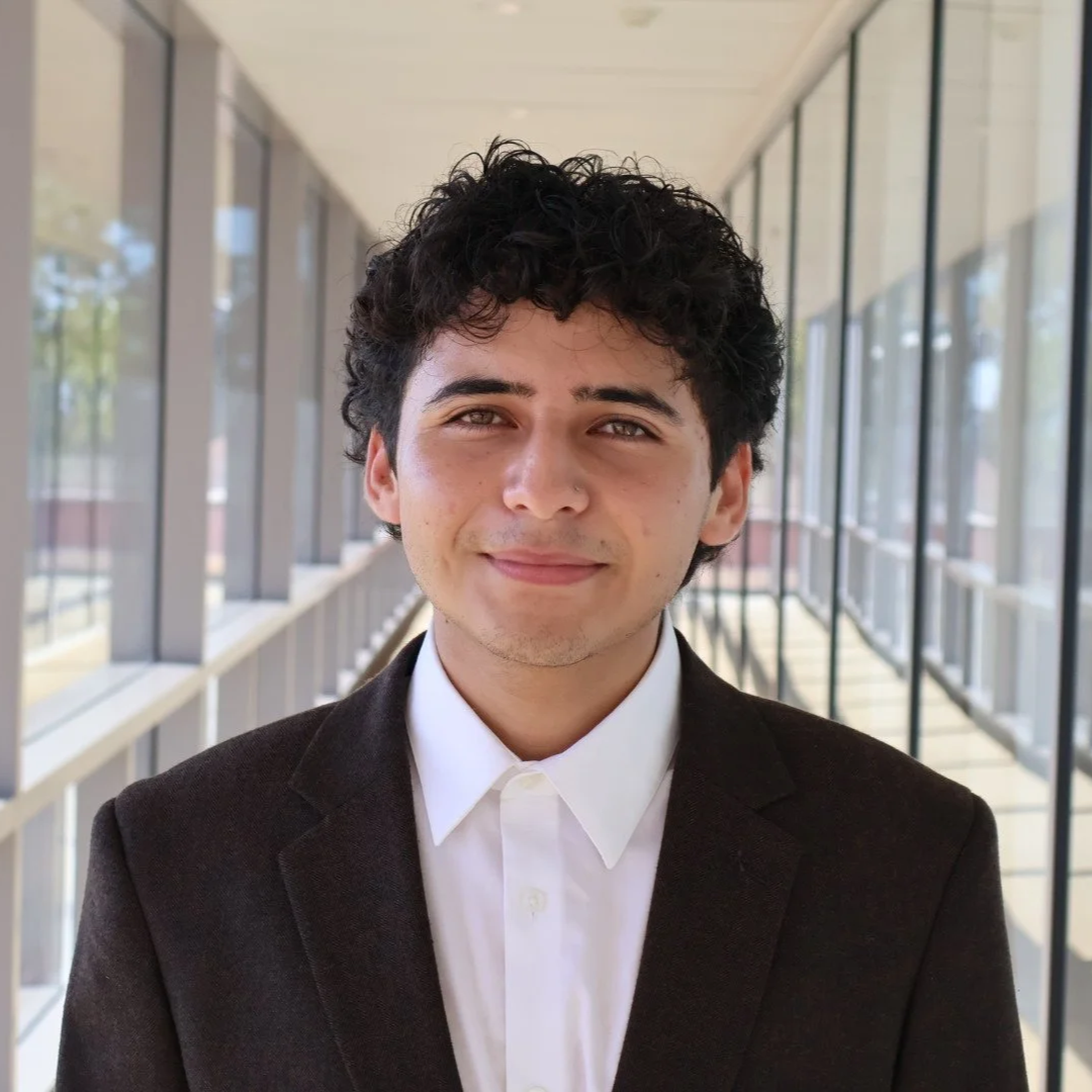 A young man with curly dark hair is wearing a blue suit and white shirt, standing outdoors with a cityscape in the background.