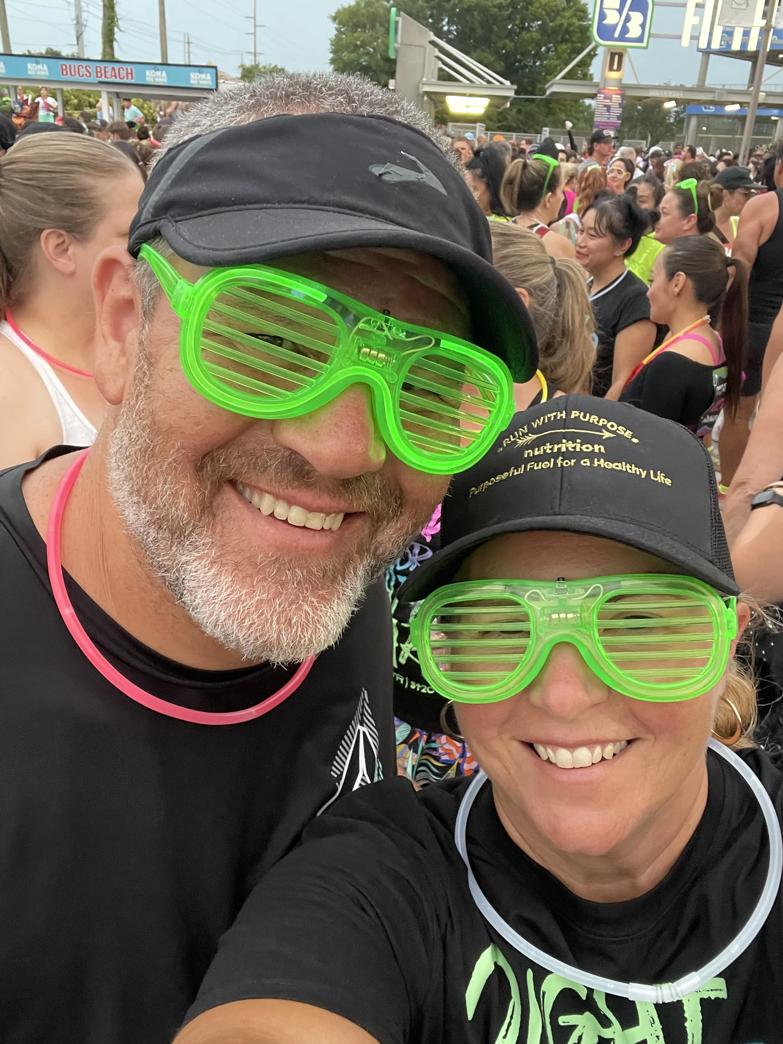 Smiling man and woman wearing neon green shutter shades, black caps, and black shirts at a crowded outdoor event with many people in the background.