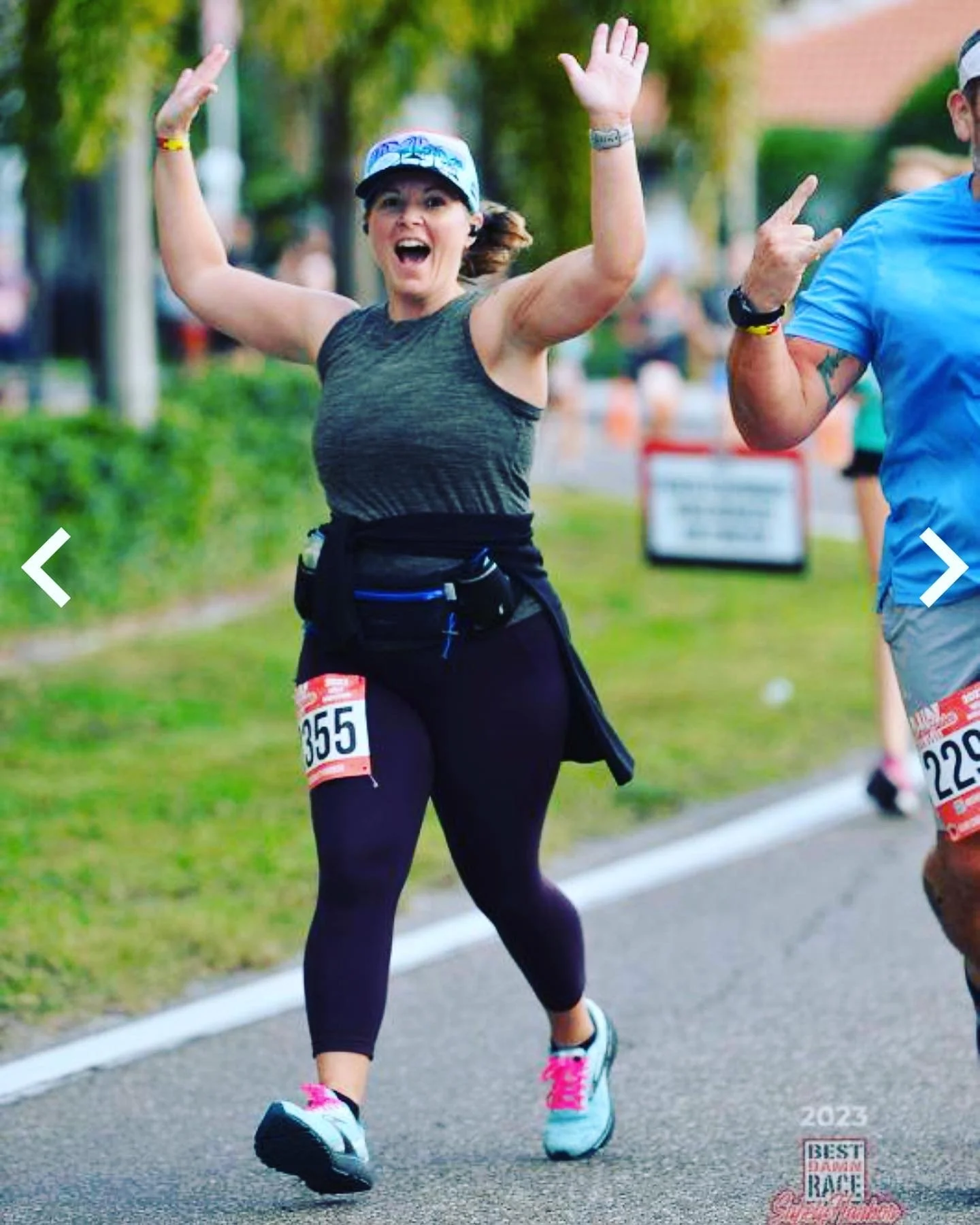 Woman smiling, raising arms, wearing a race bib number 355, participating in a marathon or race, with other runners on a paved trail outdoors.