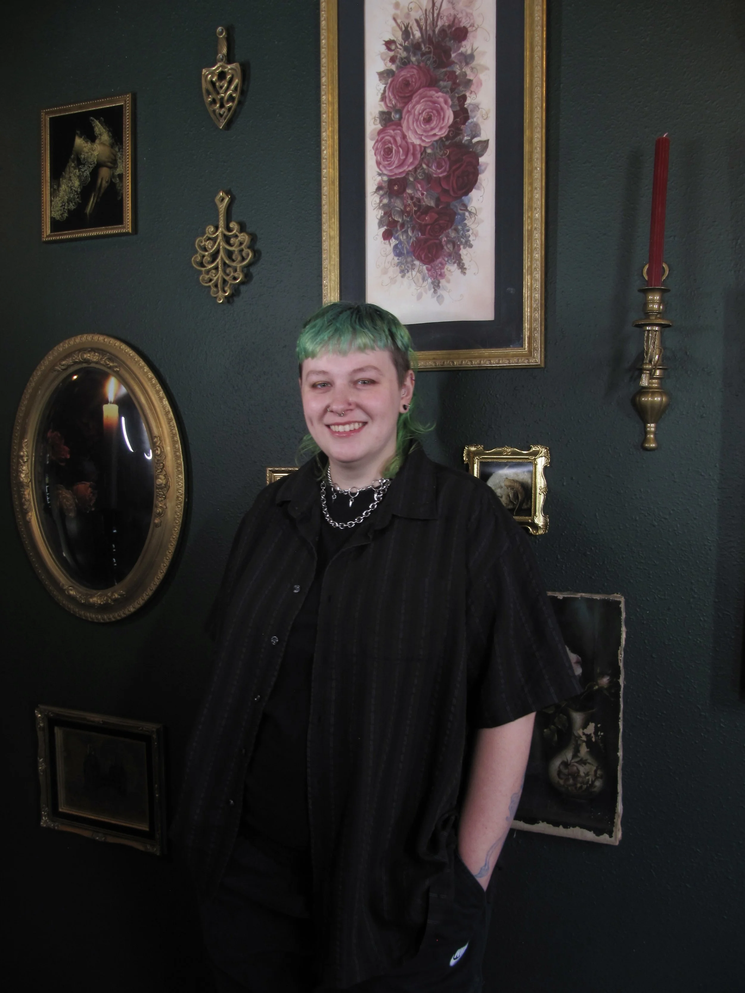 A young woman with short, multicolored hair (green, blue, and brown) smiling, wearing multiple facial piercings, a black shirt, and a necklace, sitting against a dark background.