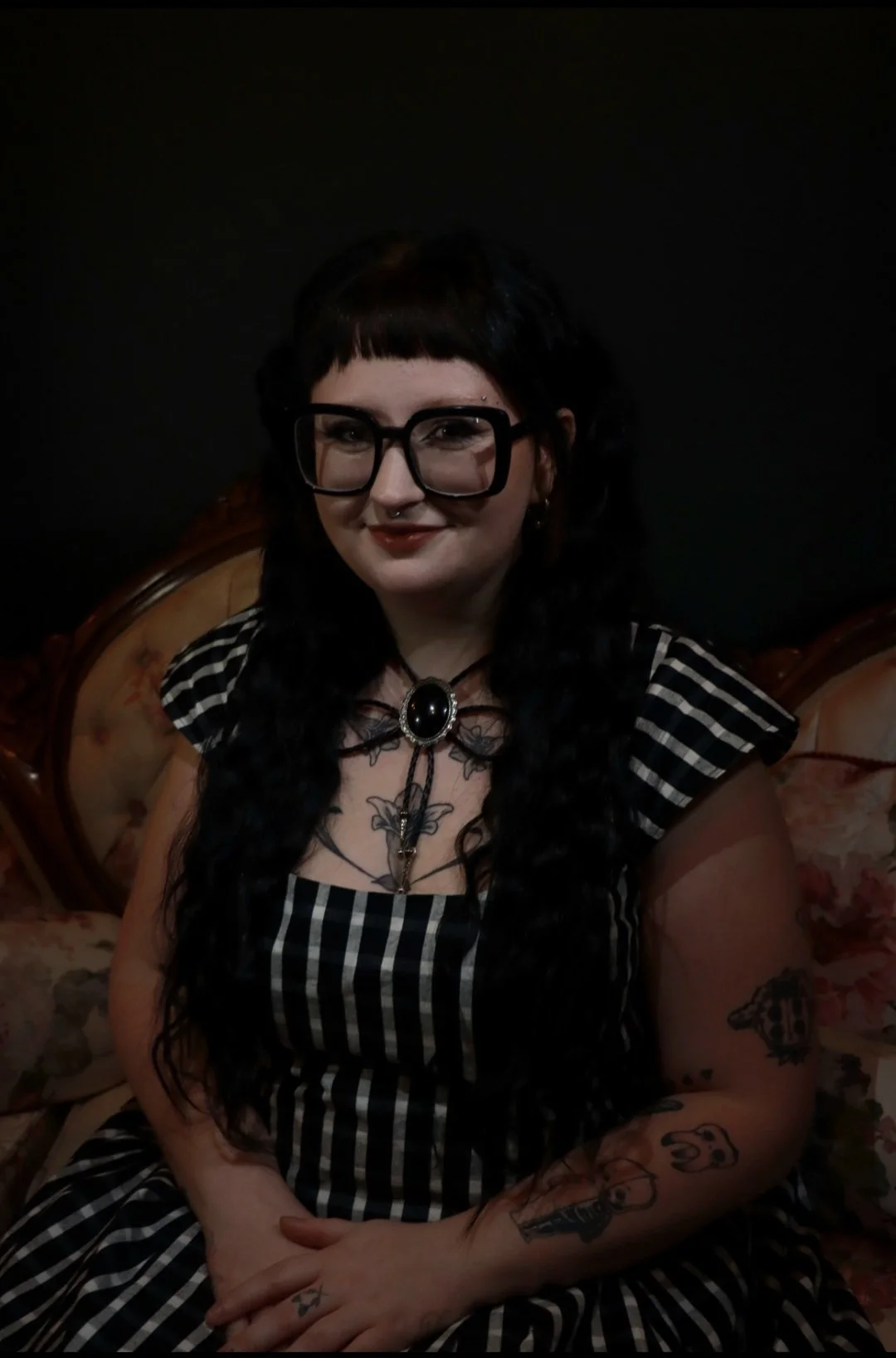 A woman with black hair and glasses, wearing a black and white striped dress, sitting on a floral vintage-style sofa, smiling at the camera.