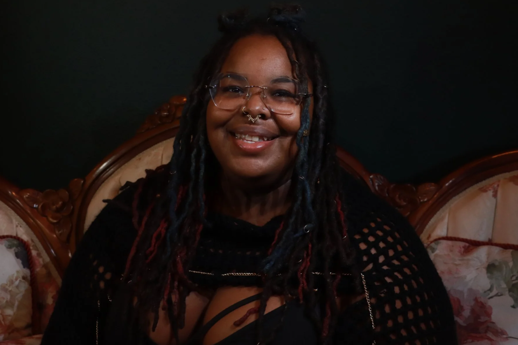 A smiling woman with dreadlocks, wearing glasses and a septum piercing, sitting on a vintage wooden sofa with floral-patterned cushions against a dark wall.