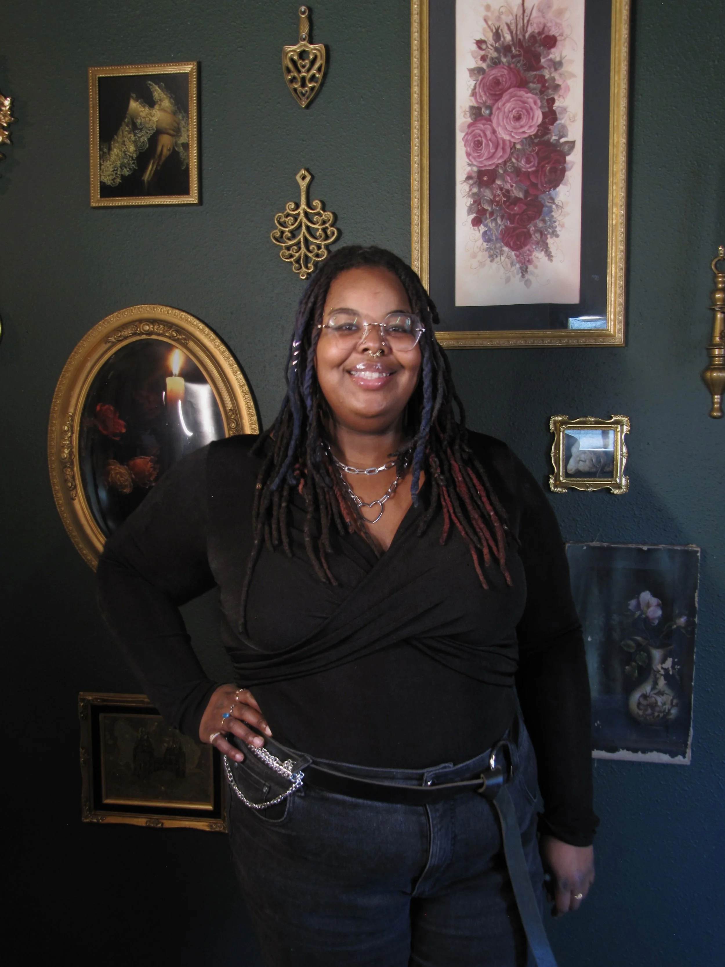 A smiling woman with dreadlocks, wearing glasses and a septum piercing, sitting on a vintage wooden sofa with floral-patterned cushions against a dark wall.