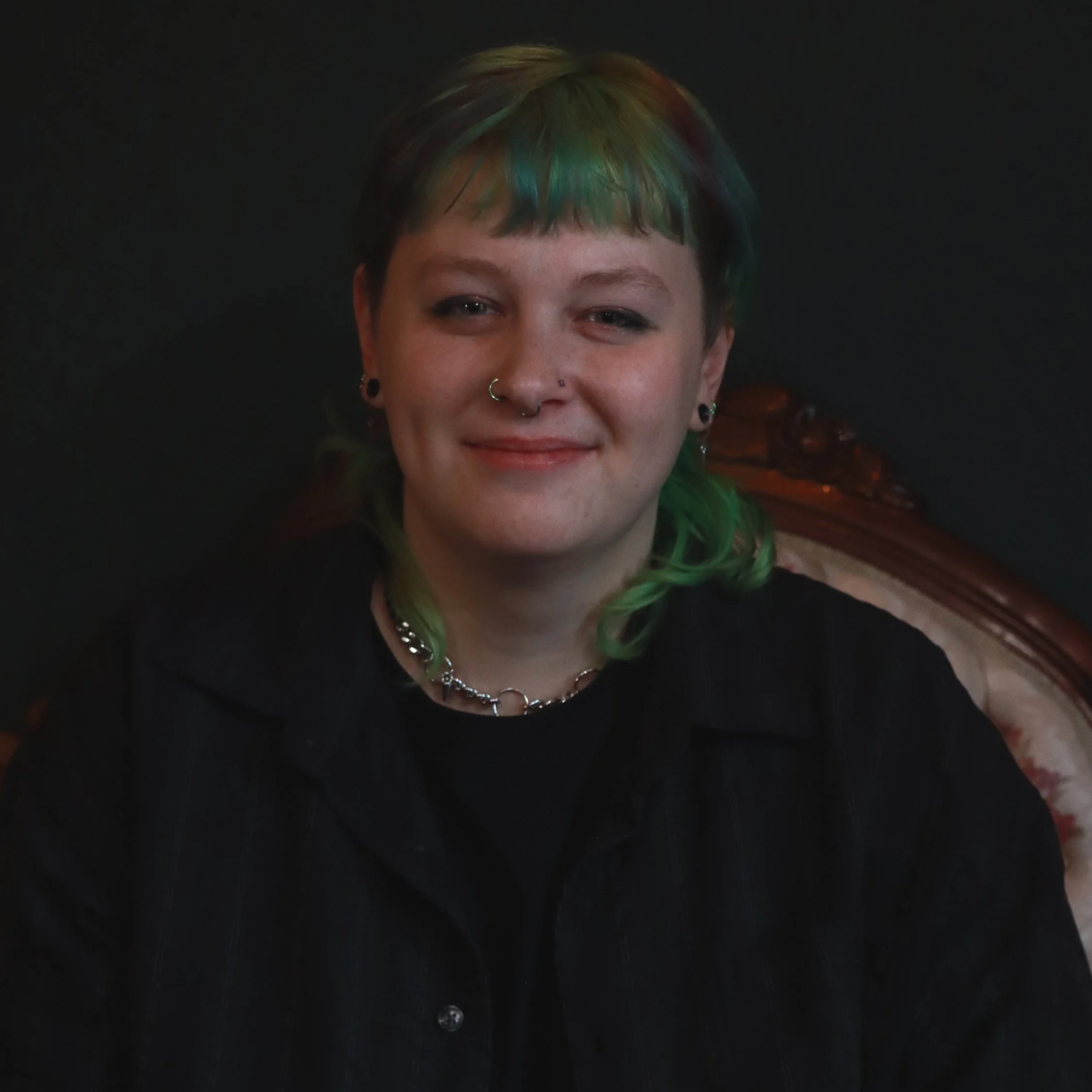 A young woman with short, multicolored hair (green, blue, and brown) smiling, wearing multiple facial piercings, a black shirt, and a necklace, sitting against a dark background.