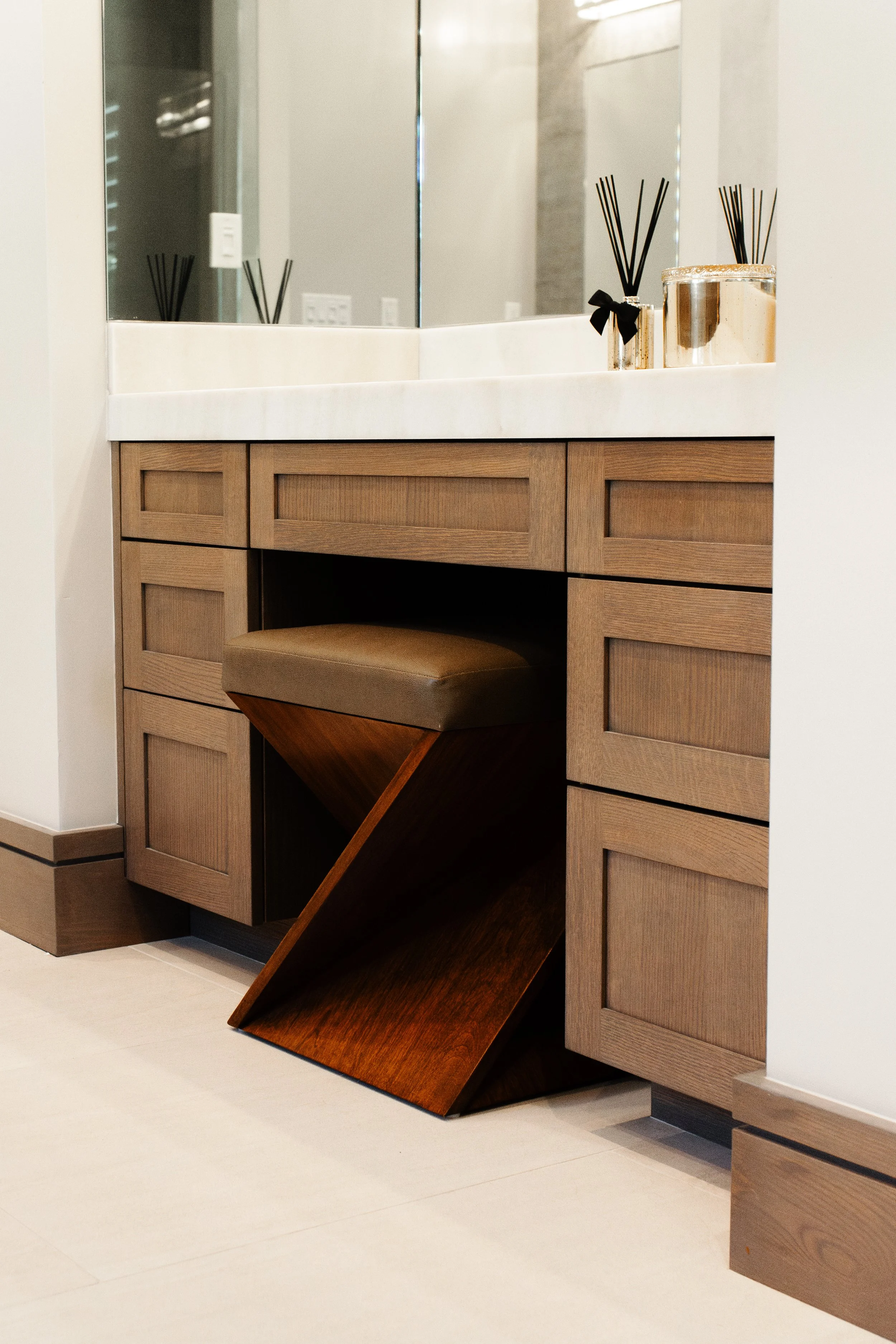 An elegant, luxurious, coastal bathroom vanity in a custom-stained wood material. The stain is a neutral, light brown, and doors are a modern shaker style. The cabinet is topped with a white stone countertop.