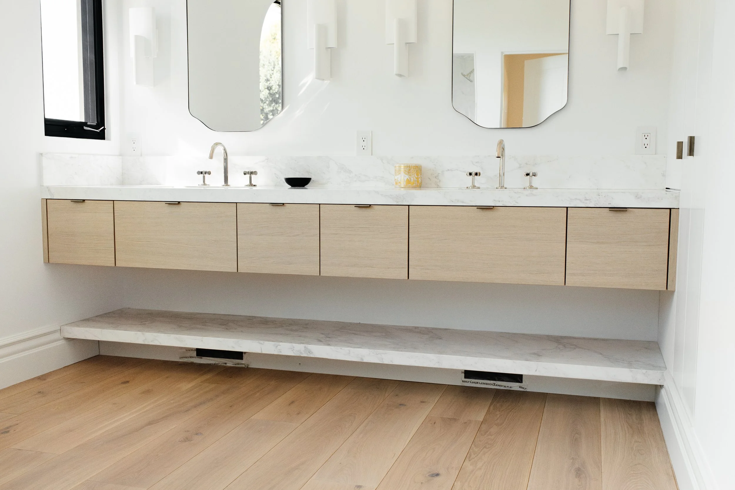 A stylish and luxurious primary bathroom vanity with dual sinks. The vanity is a floating cabinet with a marble shelf installed below. Cabinet is a modern white oak material with horizontal-grain slab drawer fronts and nickel finger pulls.