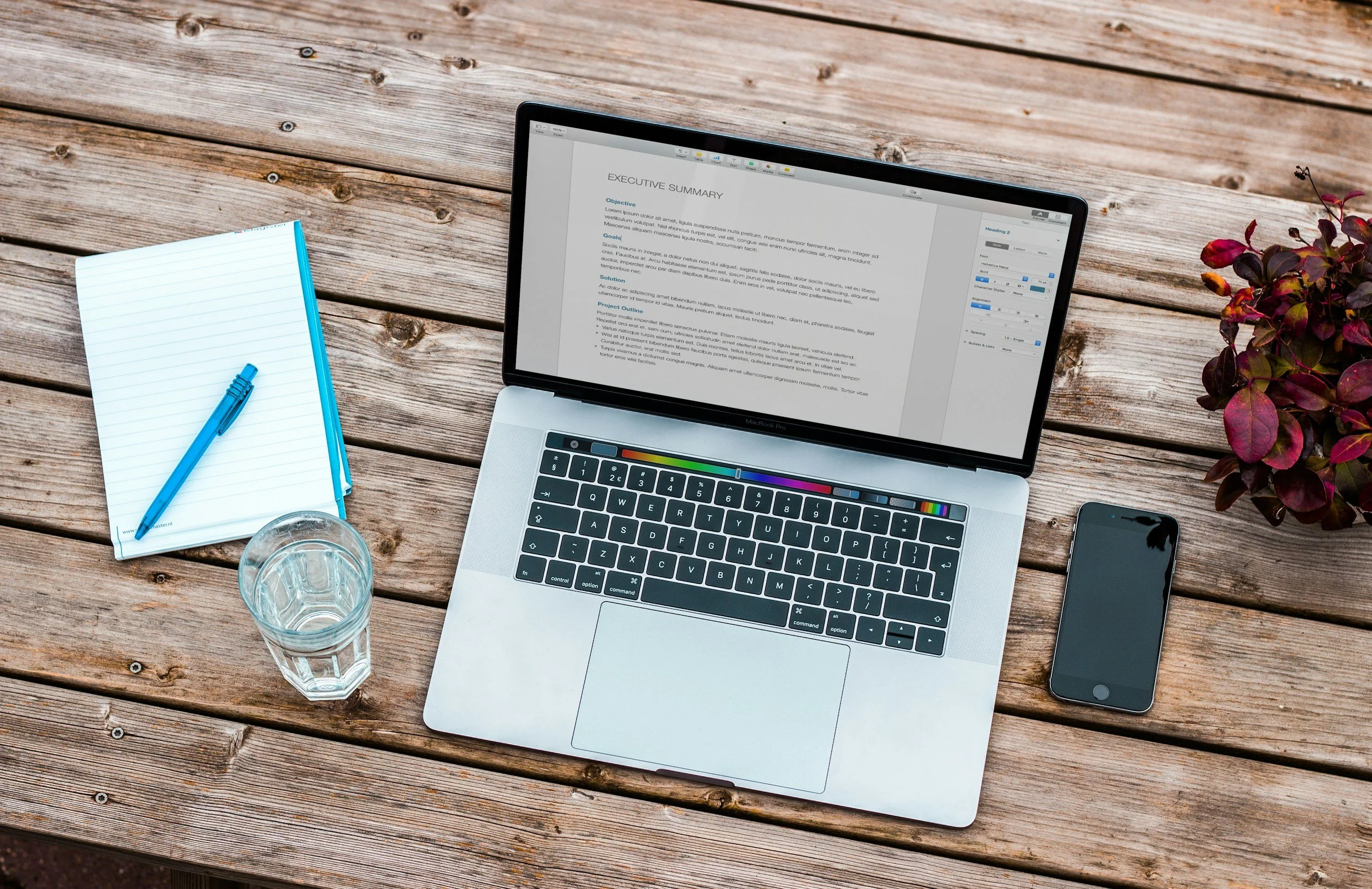 A wooden outdoor table with a silver laptop displaying a document, a black smartphone, a glass of water, a notepad with a blue pen, and a potted plant with purple and green leaves.