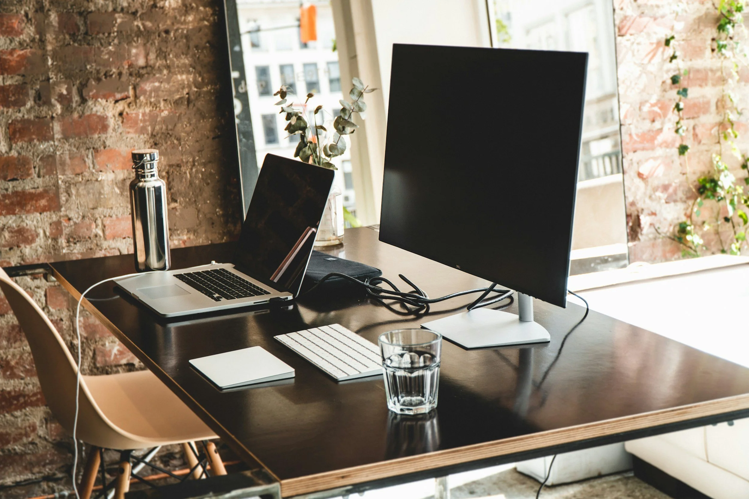 A modern workspace with a wooden table, a laptop, a desktop monitor, a glass of water, a keyboard, a notebook, and a water bottle, set against a brick wall and large windows with a city view.