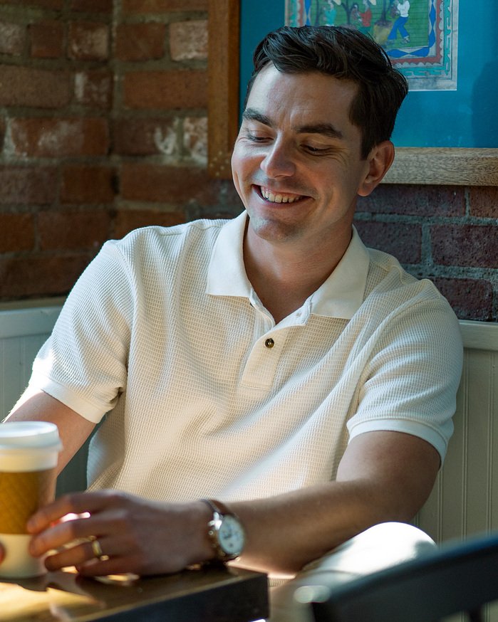 A man smiling while sitting at a table holding a cup of coffee in a cozy cafe with brick walls and colorful art in the background.