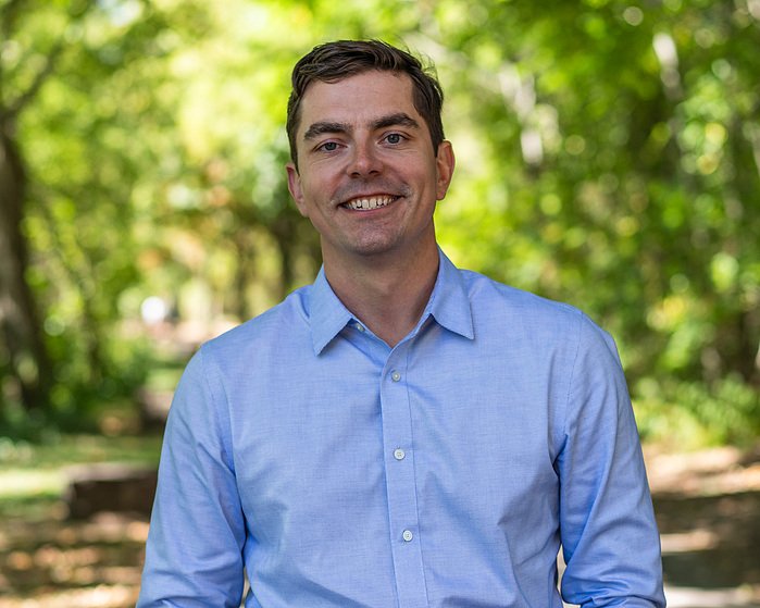 A young man with dark hair and a light blue button-up shirt, smiling outdoors with green trees in the background.