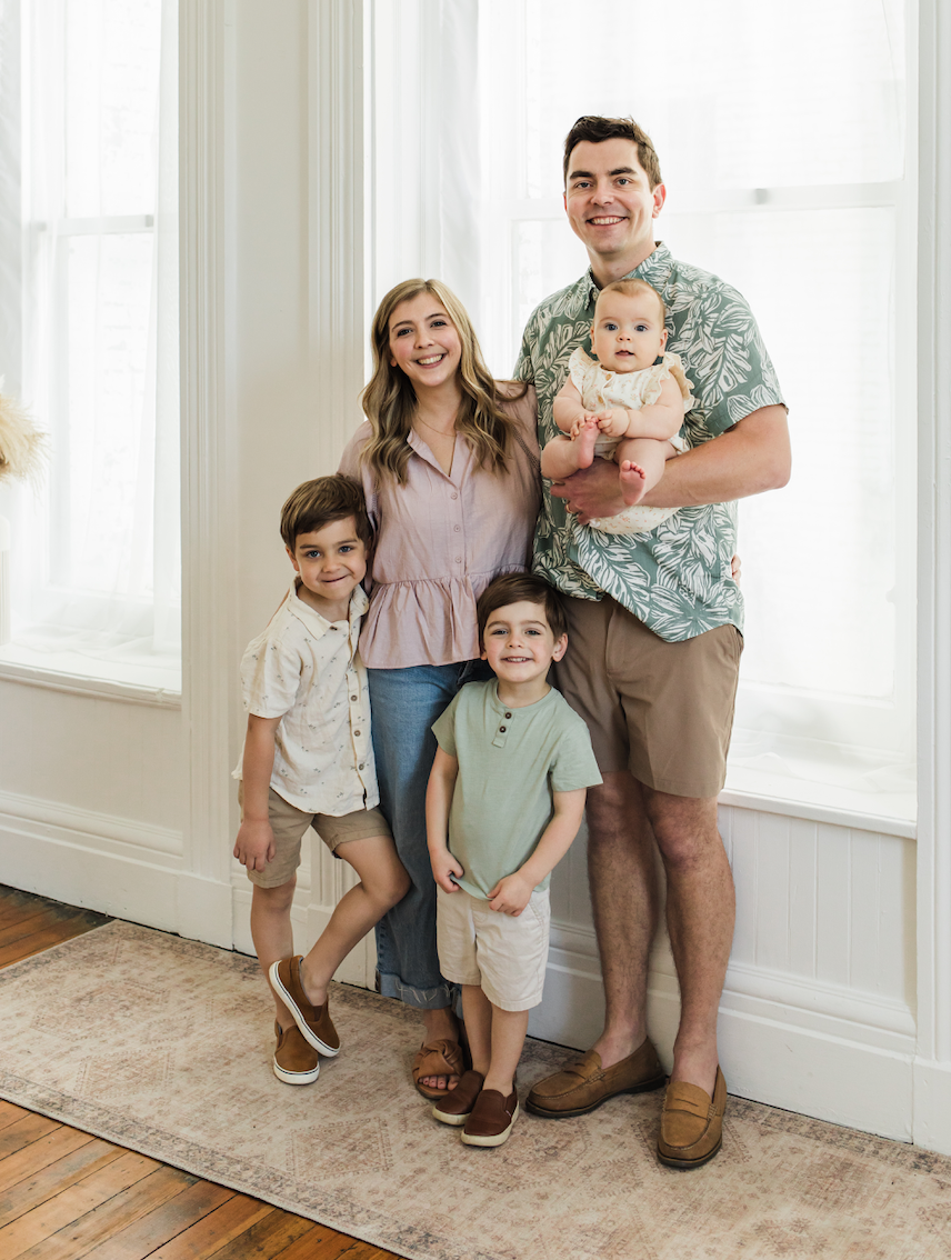 A family of five posing indoors, with large windows in the background. The father is holding a baby girl, the mother stands next to him, and two young boys stand in front of them, all smiling.