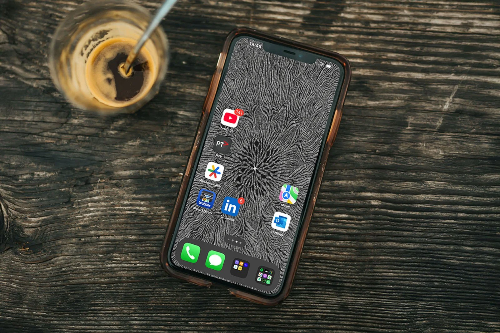 Iphone-with black-and-white line-art-wallpaper-with-homescreen-laying-on-a-rough-wooden-table-next-to-an-espresso-coffee