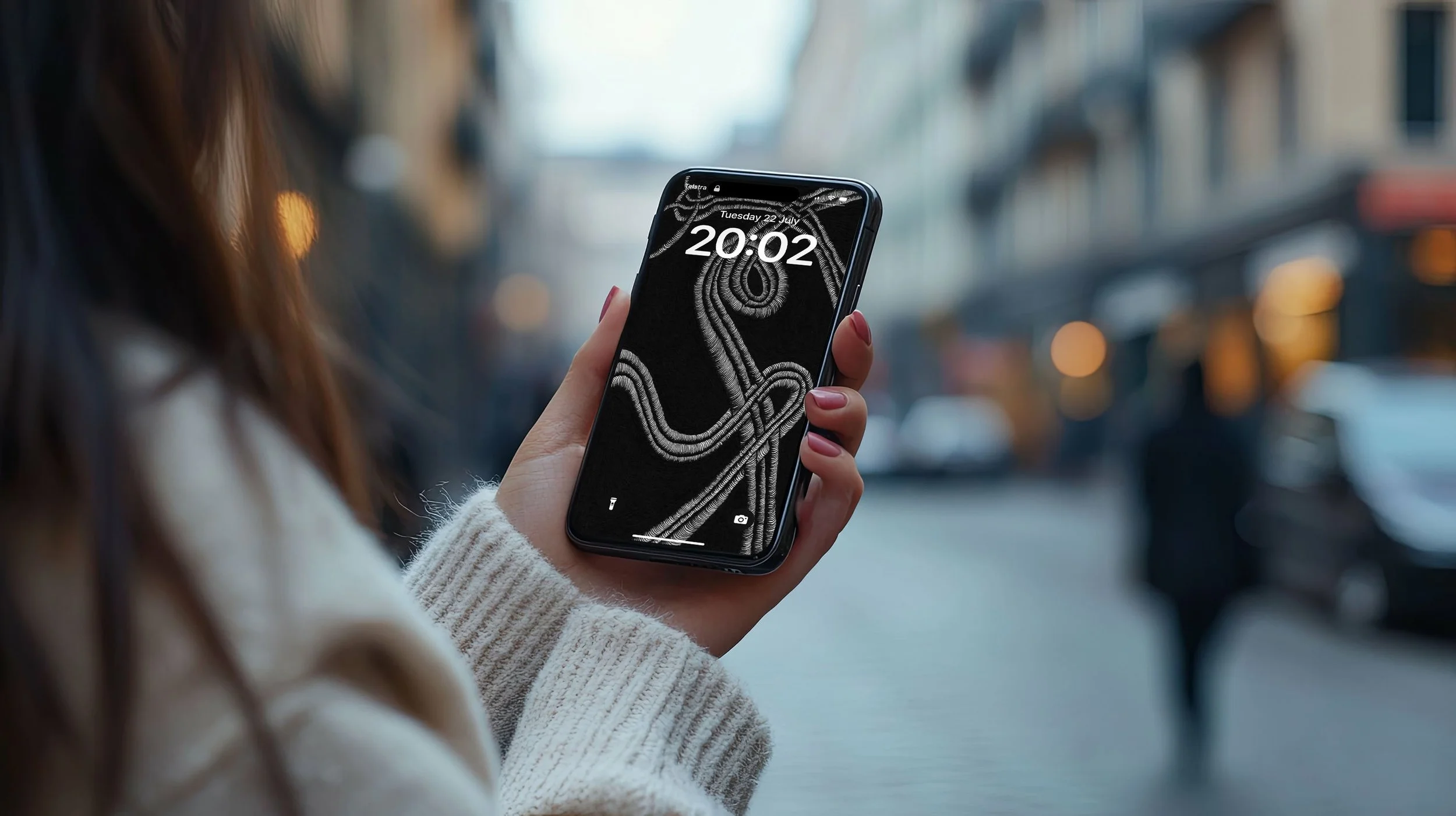 Women-with-long-brown-hair-wearing-white-sweater-standing-on-city-street-holding-an-iphone-with-two-hands-displaying-abstract-line-art-with-lockscreen.jpg