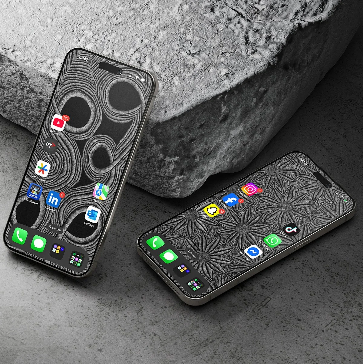 Two smartphones with patterned black and white wallpapers, placed on a concrete surface with large stone blocks in the background.