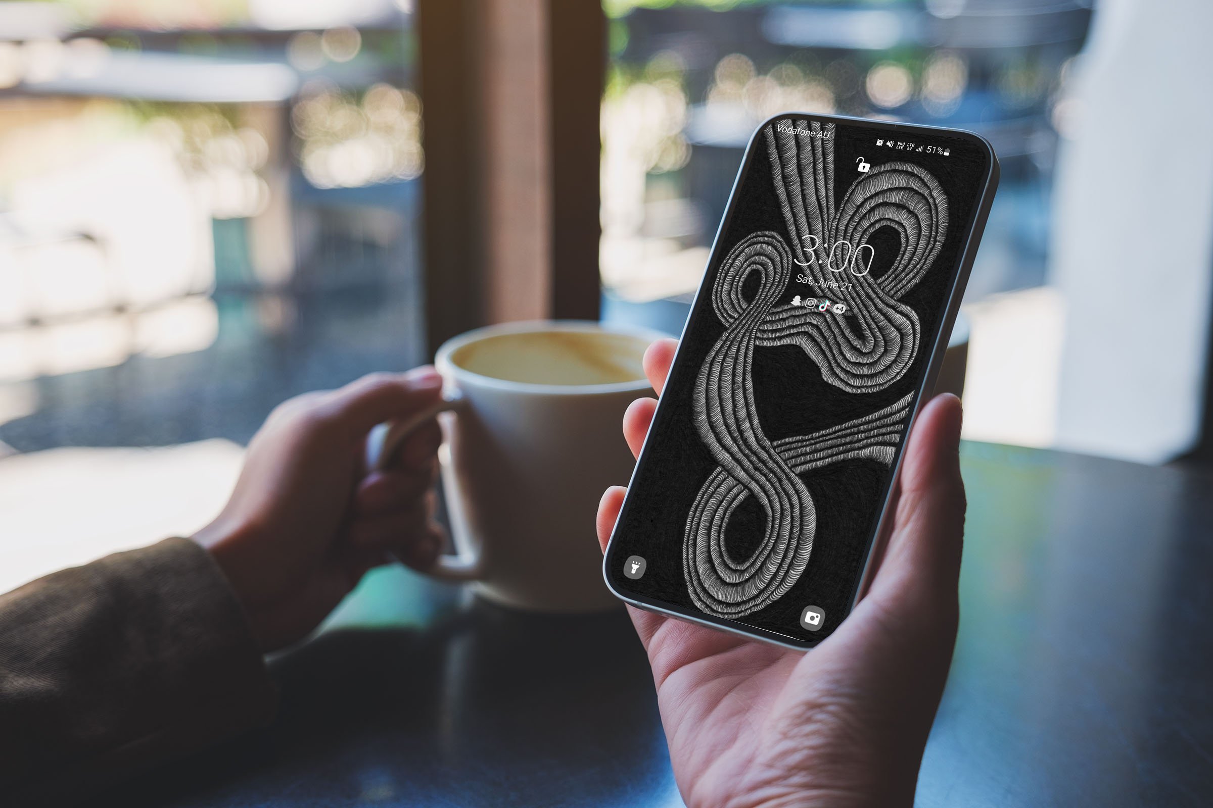 Person-sitting-holding-android-phone-with-lock-screen-and-unsonscious-flow-line-art-wallpaper-holding-a-mug-of-coffee-with-blurred-background.jpg