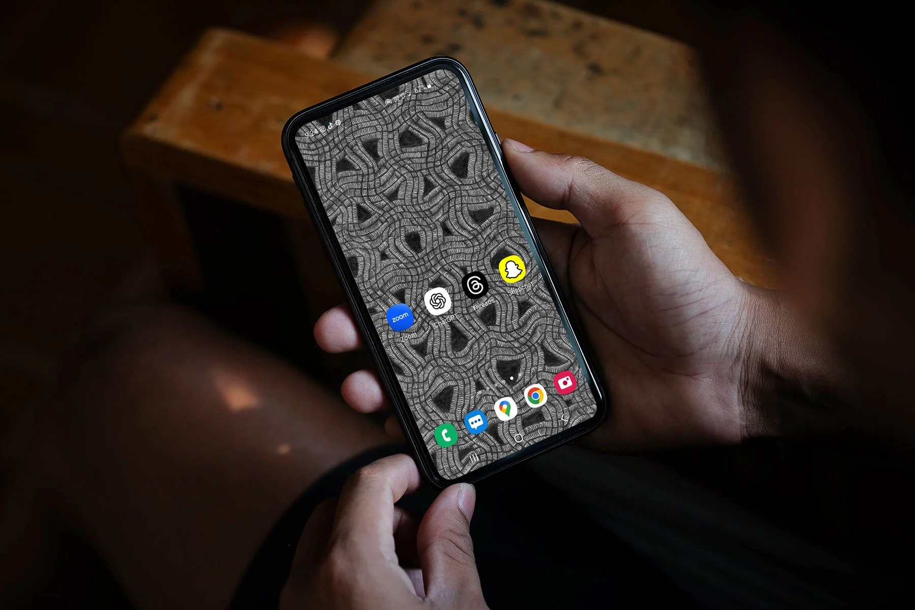 Man-sitting-holding-android-phone-with-black-and-white-line-art-wallpaper-with-app-icons-and-wooden-table in-dark-background.jpg
