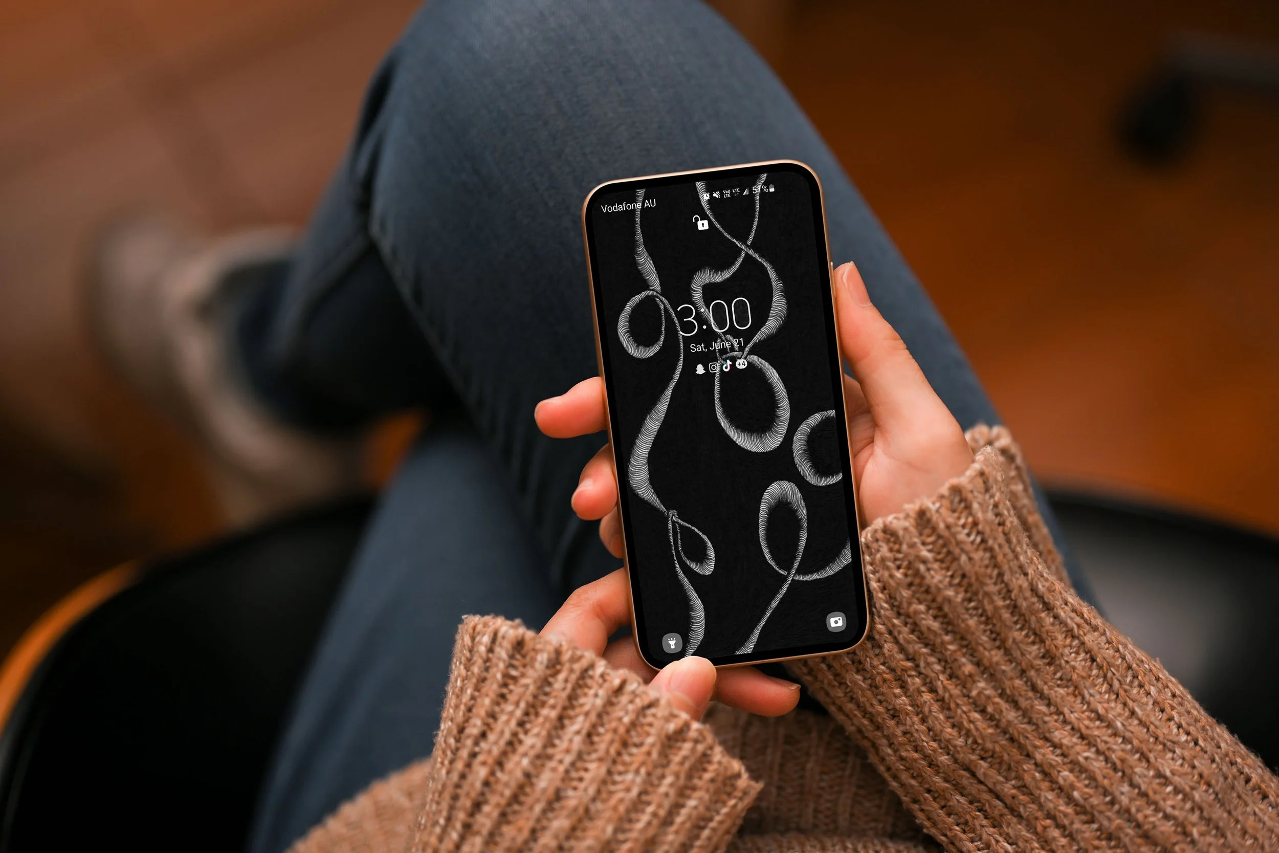 Women-sitting-wearing-a-light-brown-sweater-and-blue-jeans-holding-an-android-phone-displaying-abstract-unconscious-flow-line-art-and-lockscreen.jpg