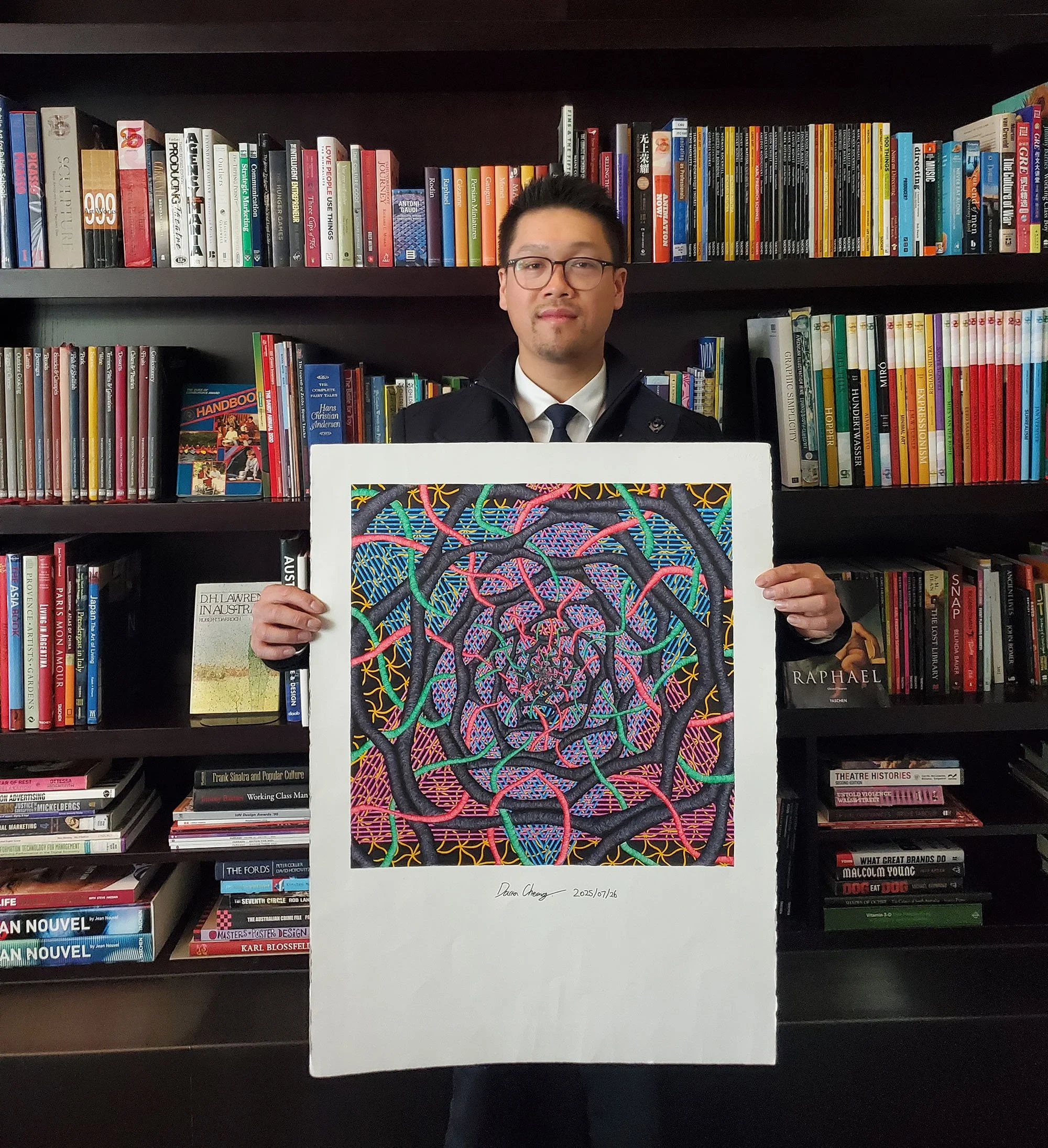 A man in glasses and a suit holding a colorful abstract artwork in front of a bookshelf filled with books.