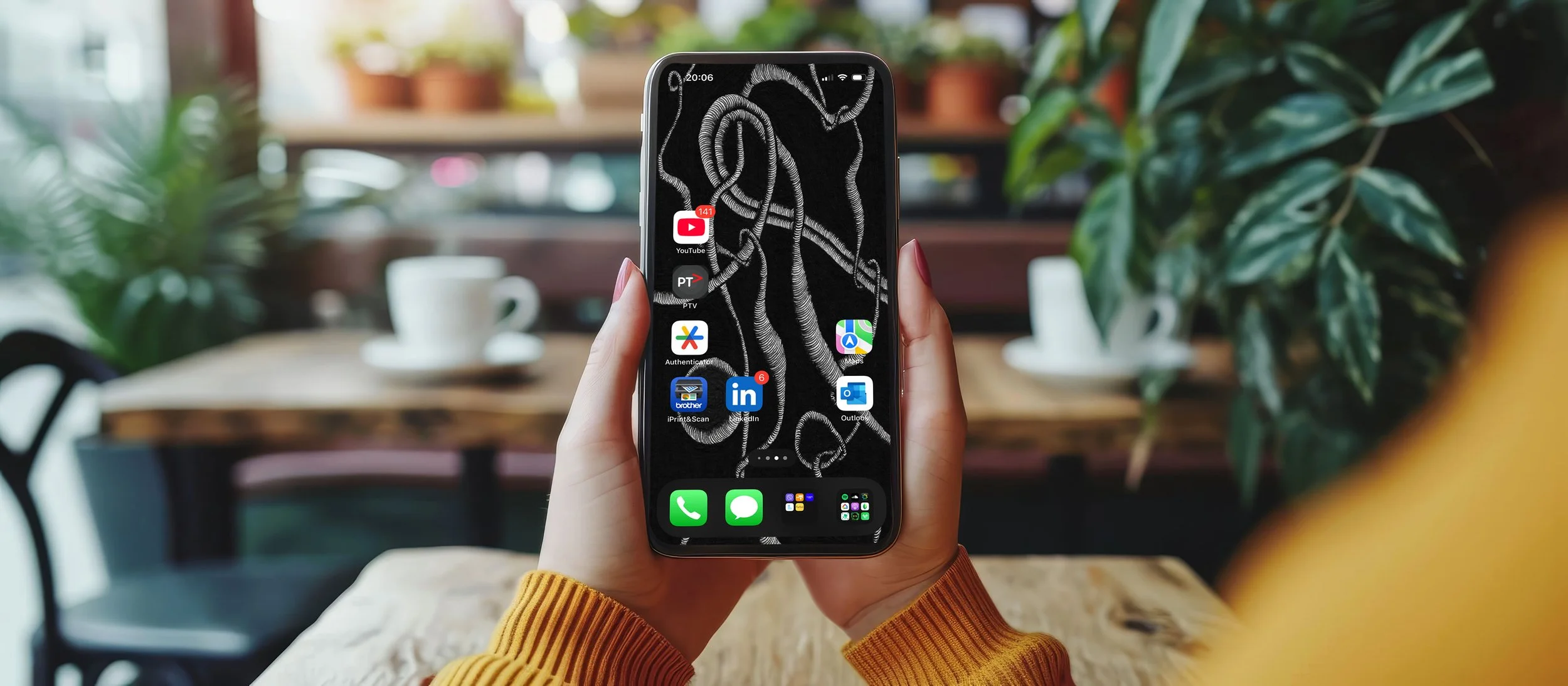Women-wearing-a-yellow-sweater-sitting-in-a-cafe-with-indoor-plants-holding-an-apple-phone-with-two-hands-displaying-a-homescreen-witjh-app-icons.jpg