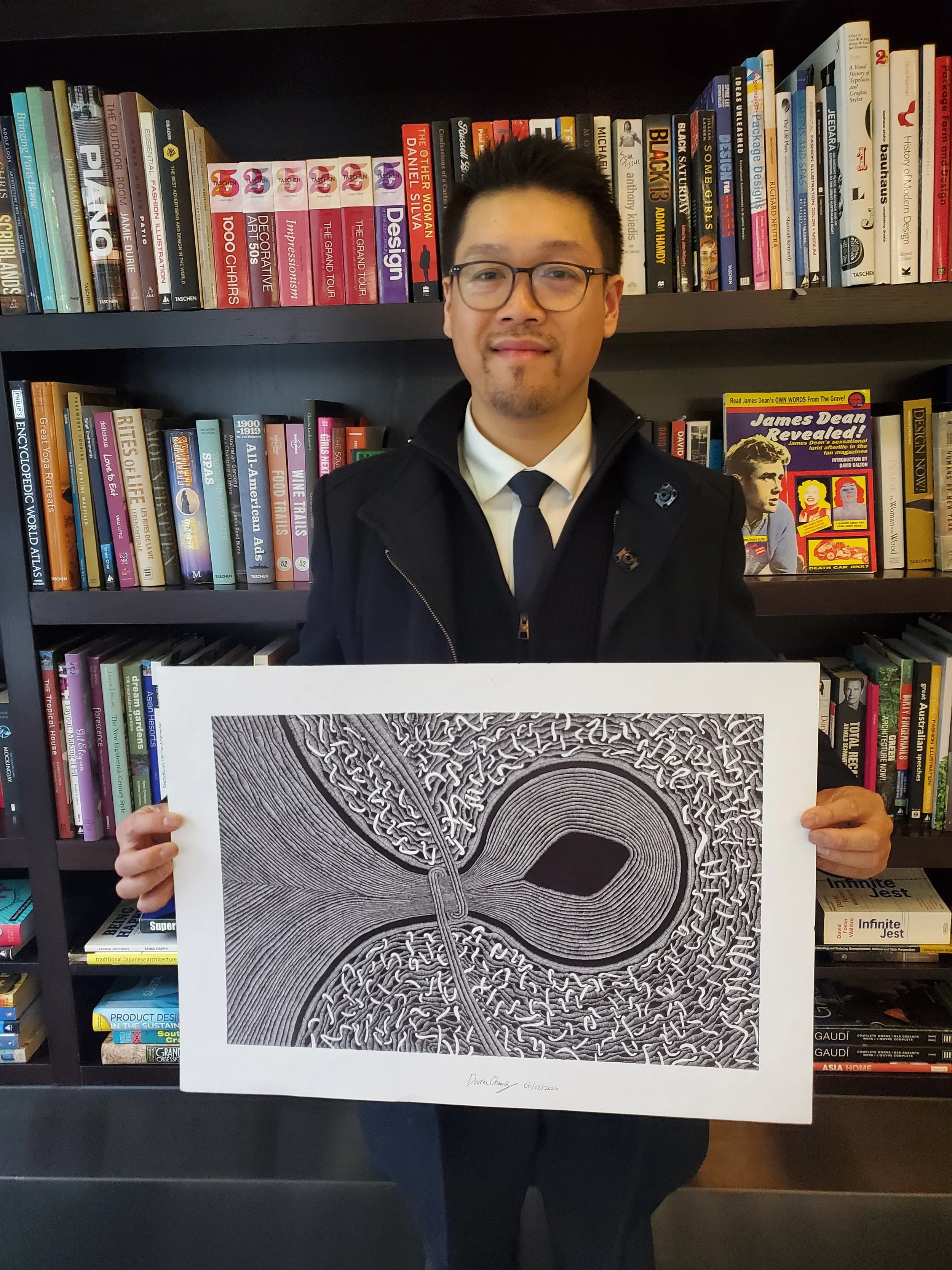A man in glasses, a white shirt, and a black coat holding a black and white abstract art print in front of a bookshelf filled with colorful books.