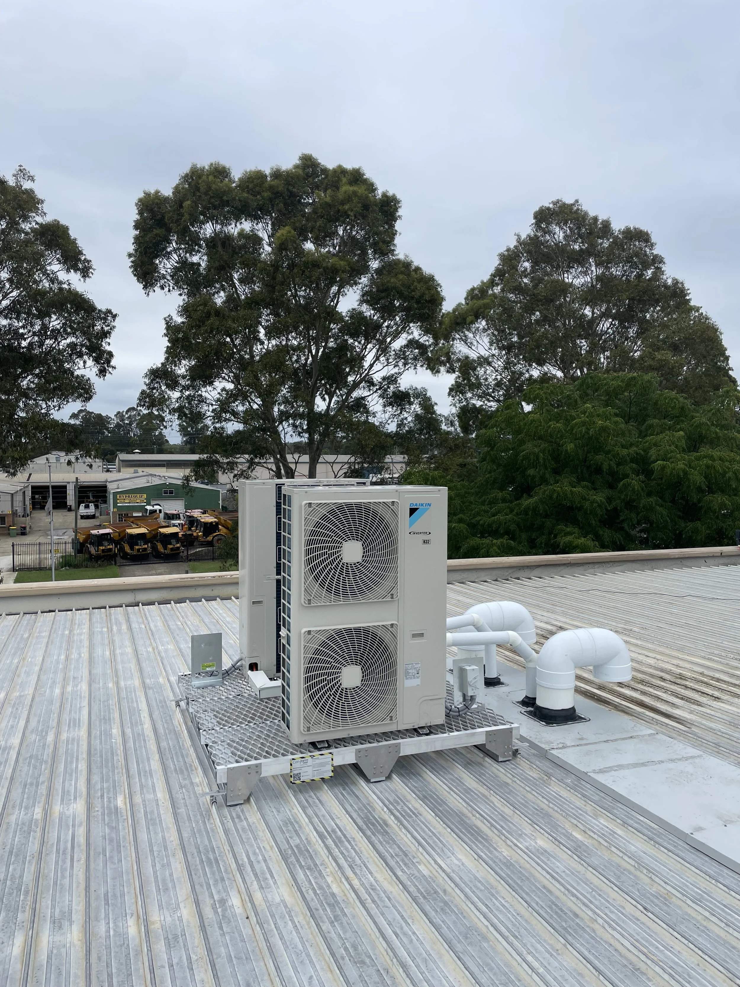 Rooftop HVAC unit with pipes and ducts, surrounded by trees and an industrial area in the background.