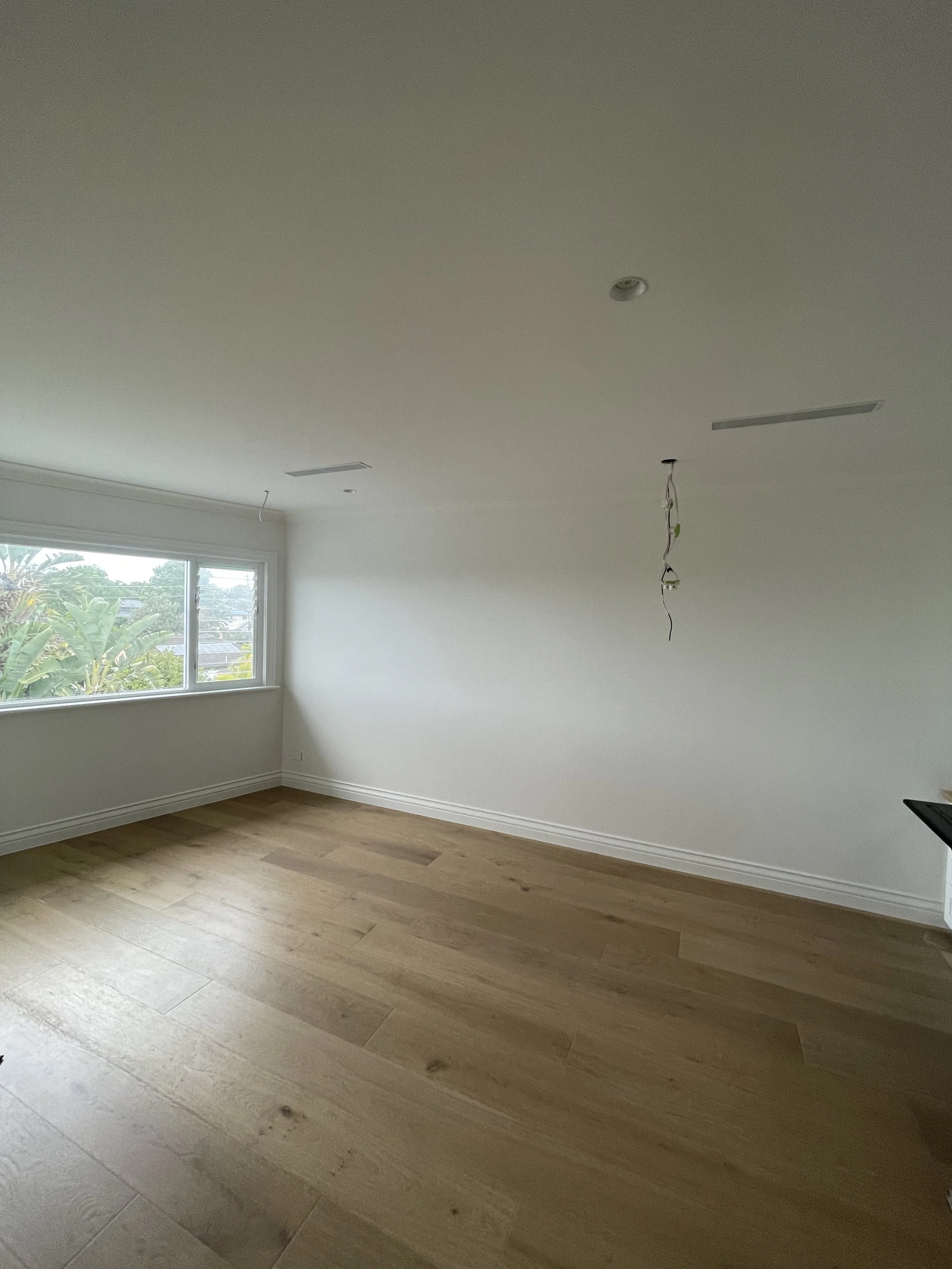 Empty room with white walls, wooden flooring, large window showing trees outside, and electrical wires hanging from the ceiling.