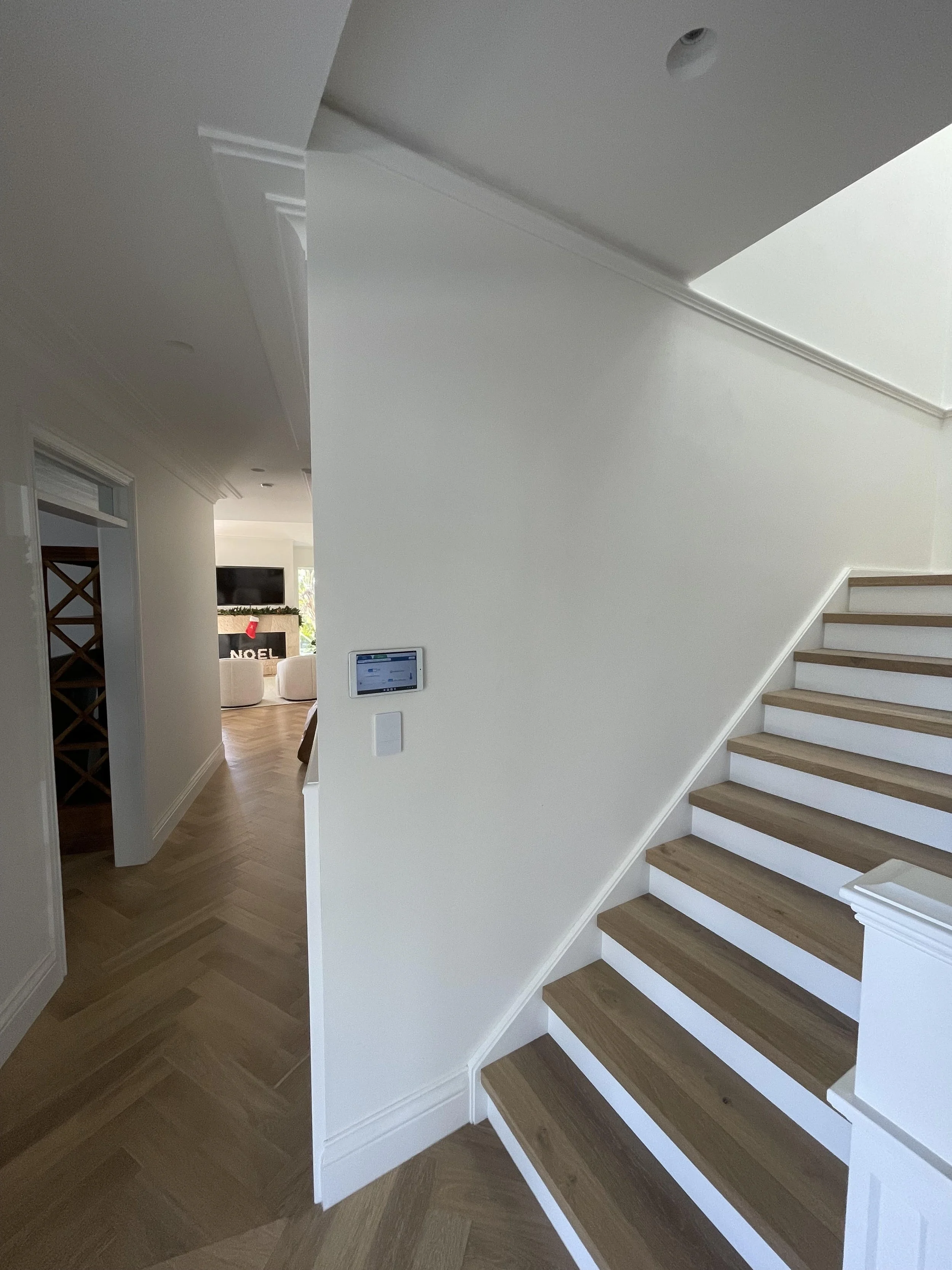 Interior of a modern home with wooden staircase, white walls, and a partial view of a living room with a TV and holiday decorations.