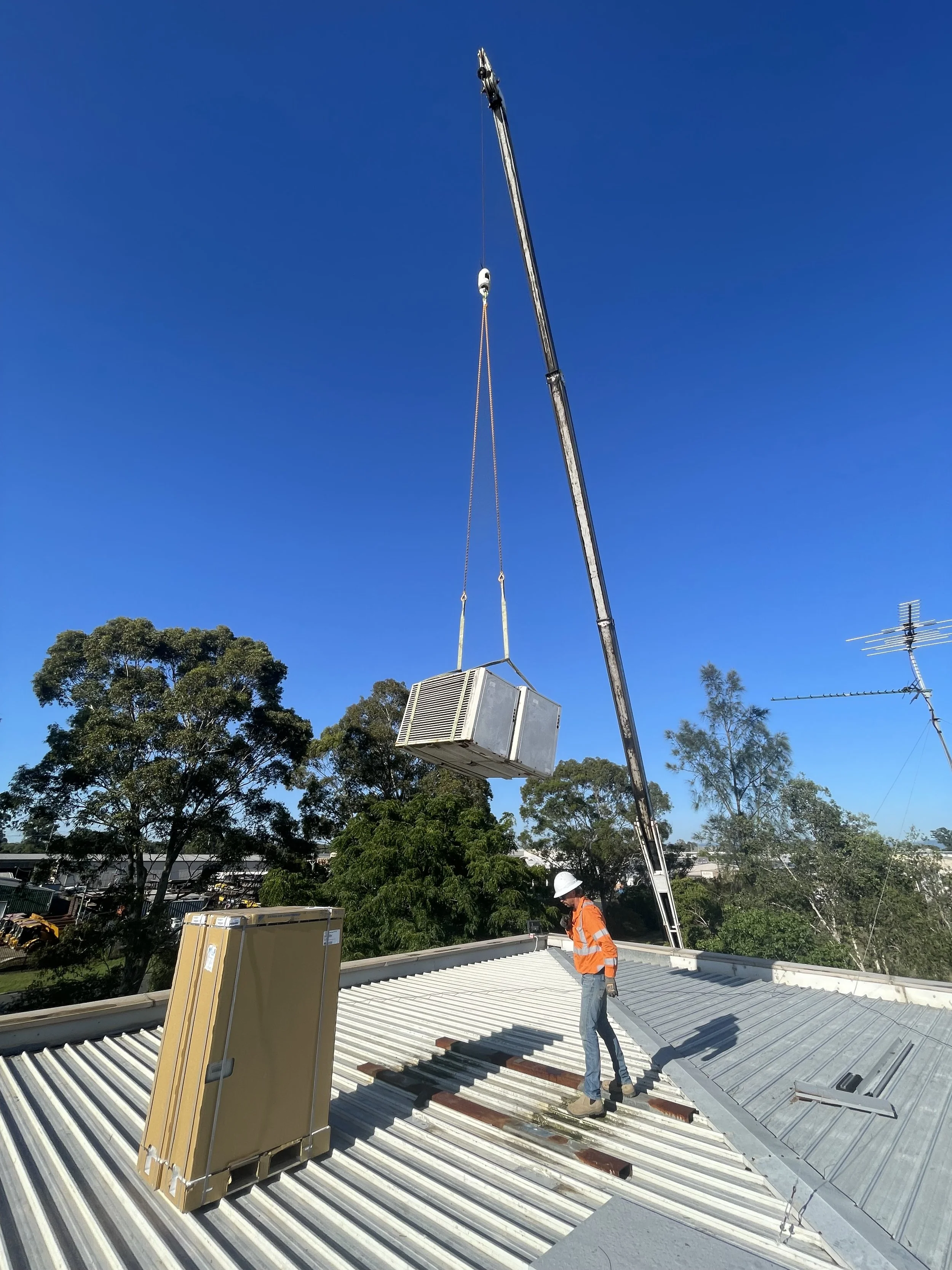 A construction worker in a safety helmet and orange safety vest standing on a metal roof as a crane lifts a large air conditioning unit into place, with trees and antennas in the background.