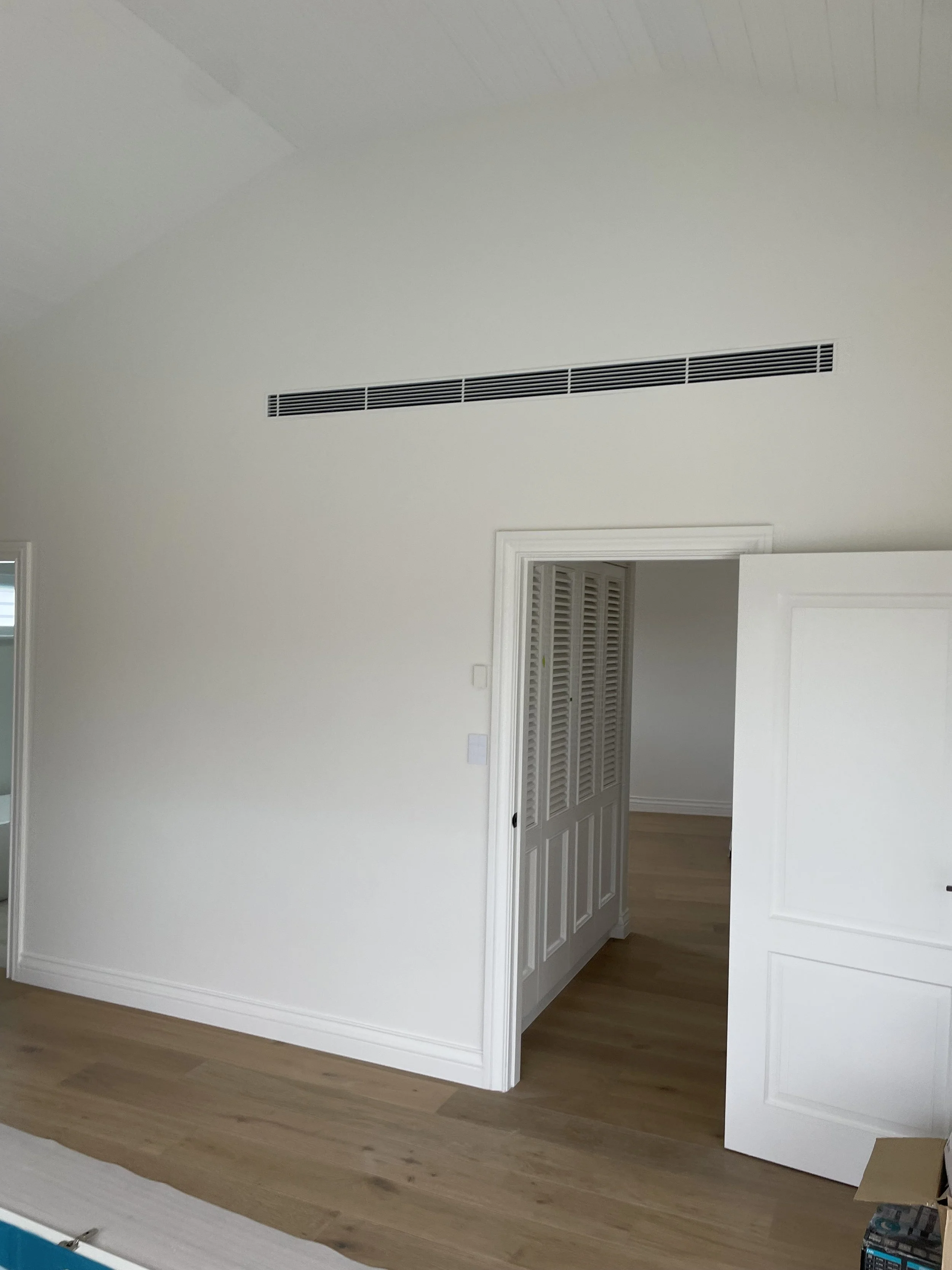 An empty room with white walls, a wooden floor, an air vent high on the wall, and an open door leading to a closet or another room.