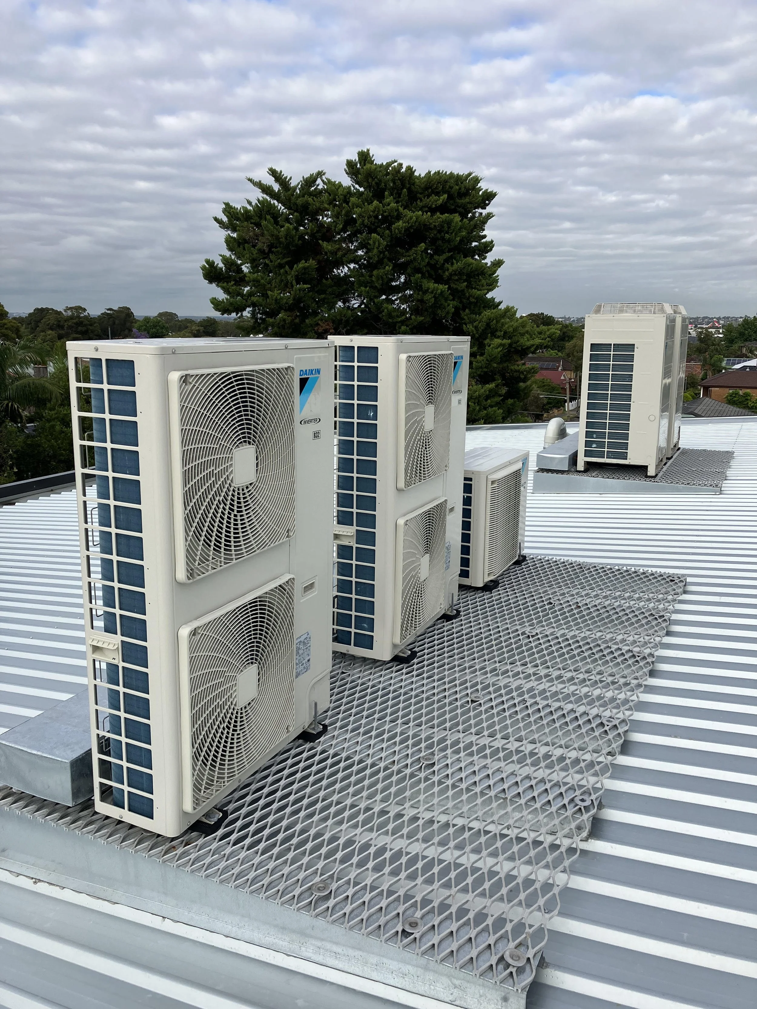 Multiple outdoor air conditioning units installed on a metal rooftop, with a tree and cloudy sky in the background.