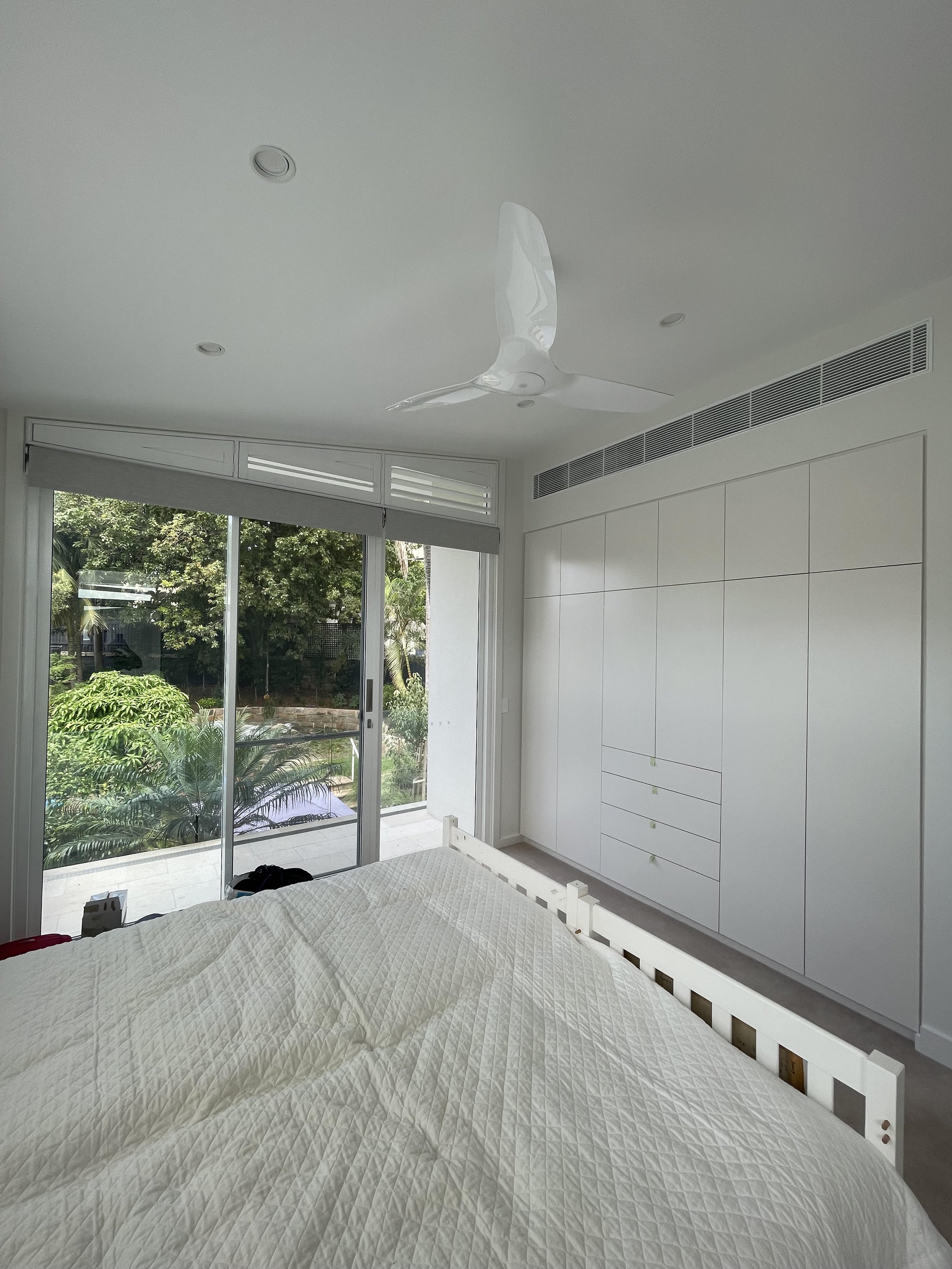 A bright, modern bedroom with a standing bed, a ceiling fan, large glass sliding doors leading to a balcony, and a white built-in wardrobe.