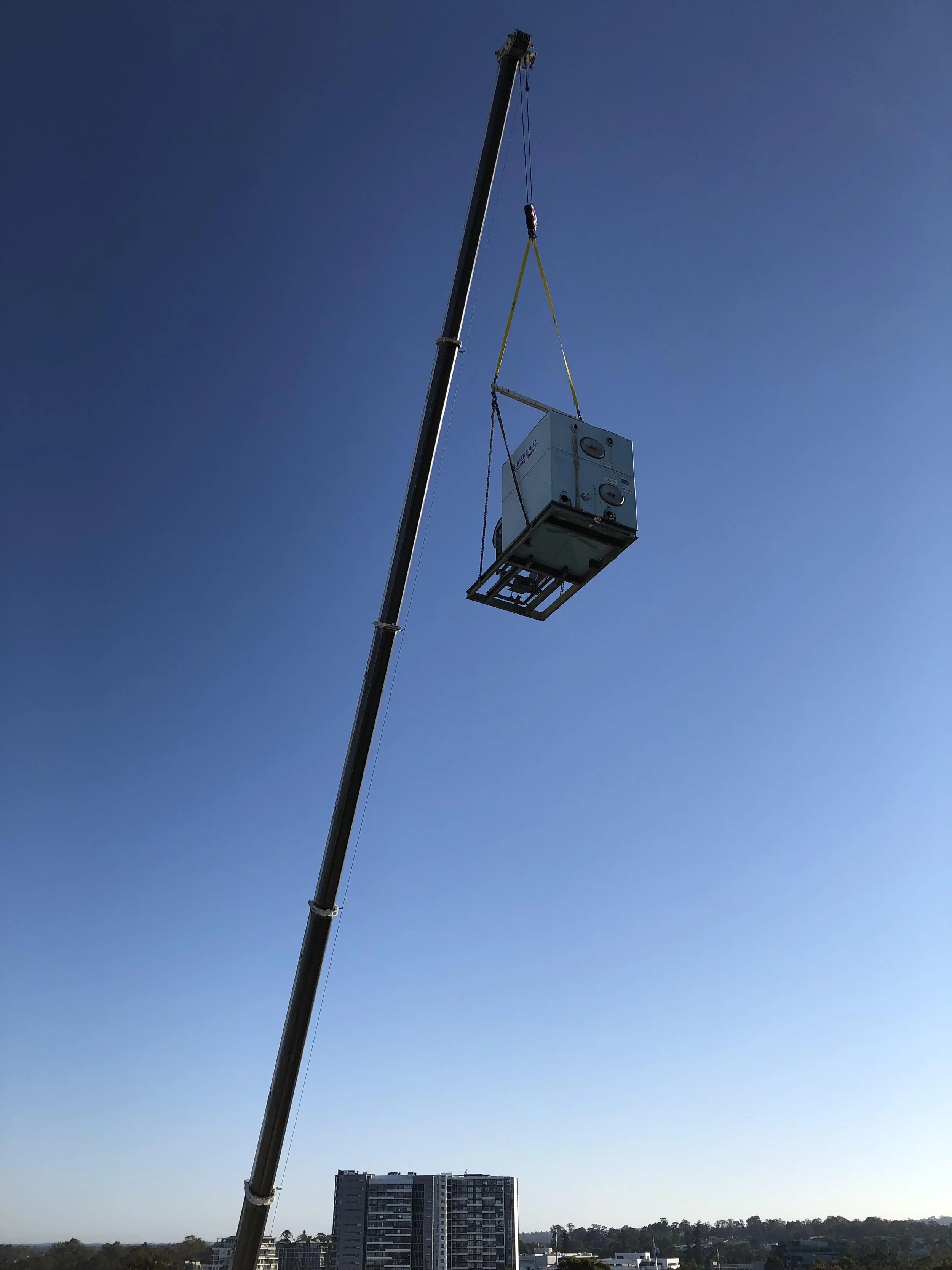 A crane lifting a large appliance, possibly an air conditioning unit, above a cityscape with high-rise buildings and trees in the background.