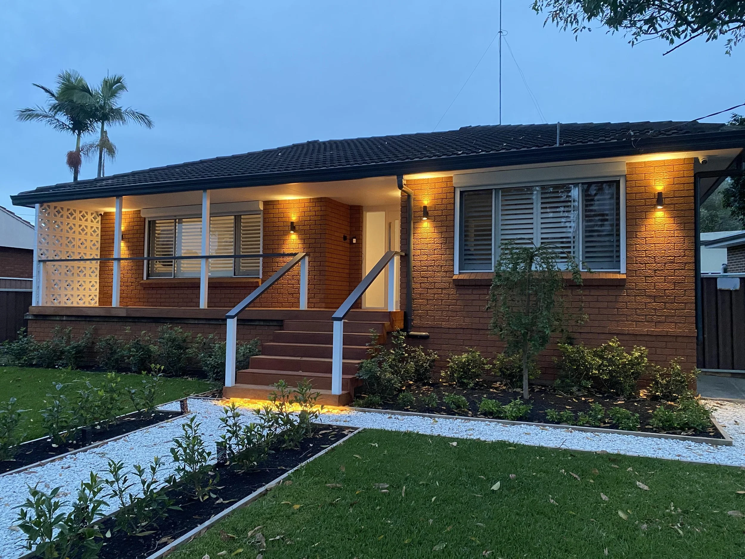 A brick house with a front porch and steps, illuminated by outdoor wall lights, with a garden and white gravel path in the front yard during dusk.