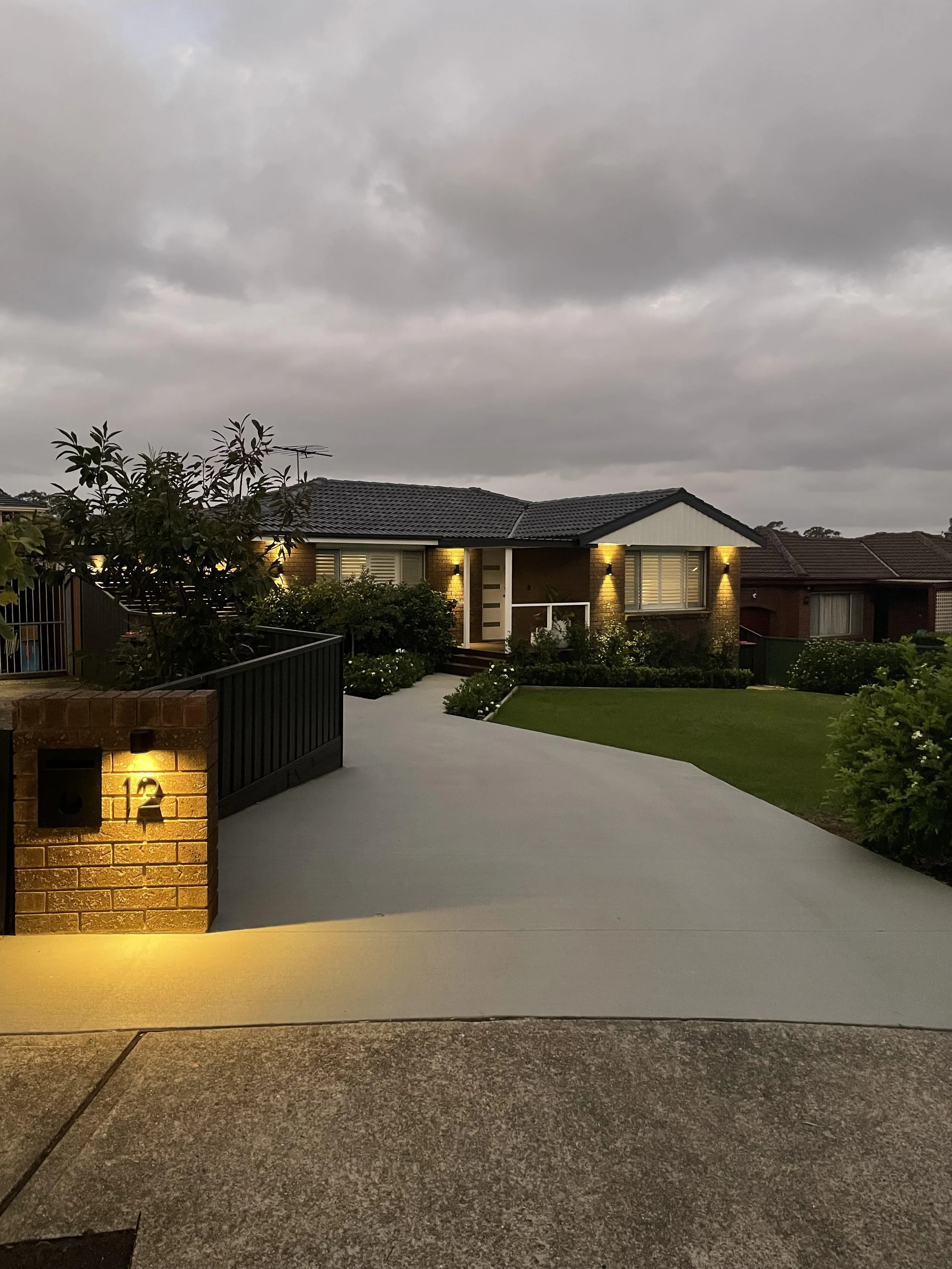 Front view of a single-story house at dusk with modern exterior lighting, a concrete driveway leading to the house, green lawn, and landscaping beneath a cloudy sky.