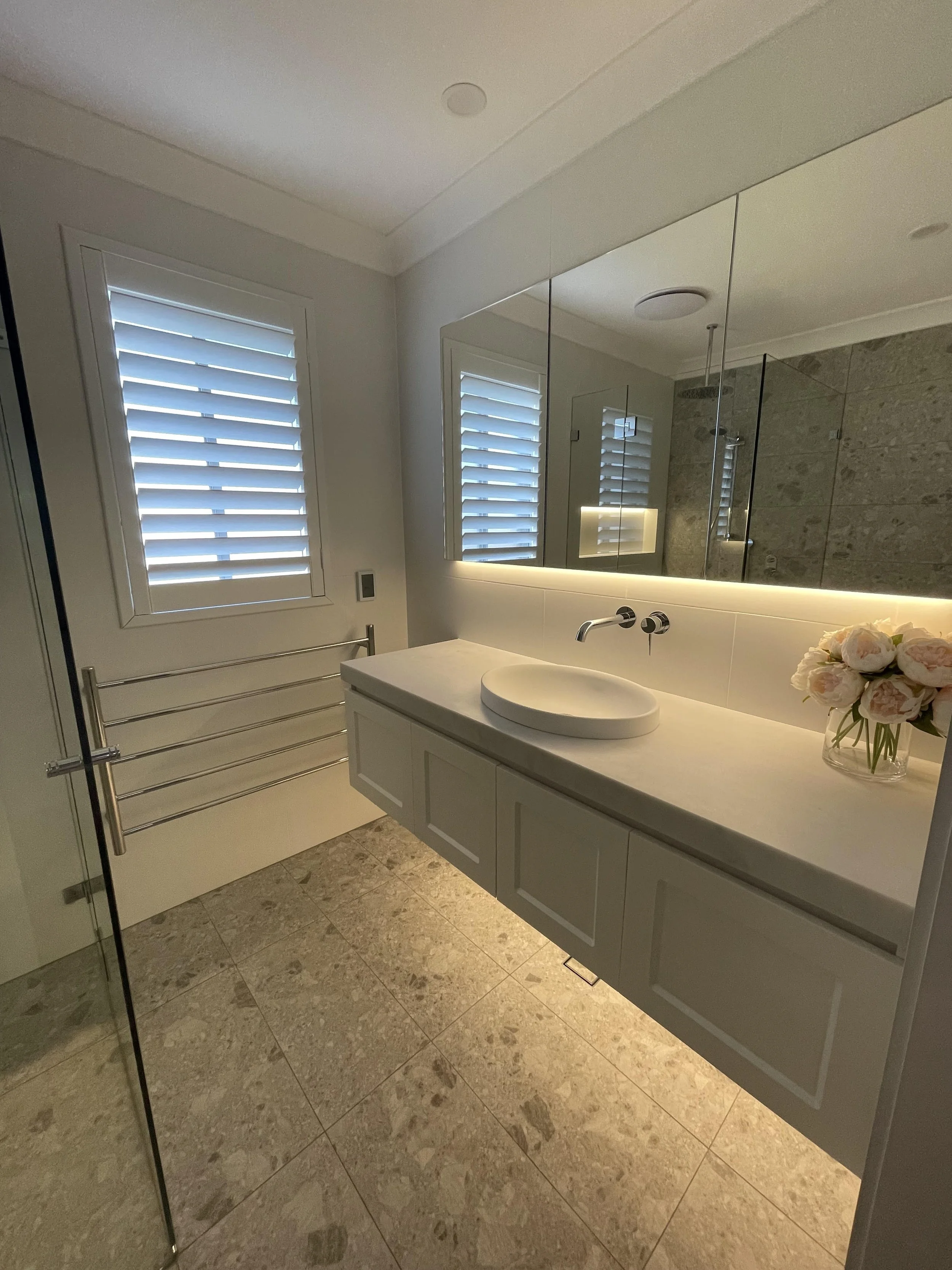 Modern bathroom with a floating vanity, white basin sink, large mirror, window with plantation shutters, floral arrangement, and walk-in shower with glass enclosure and stone tiling.