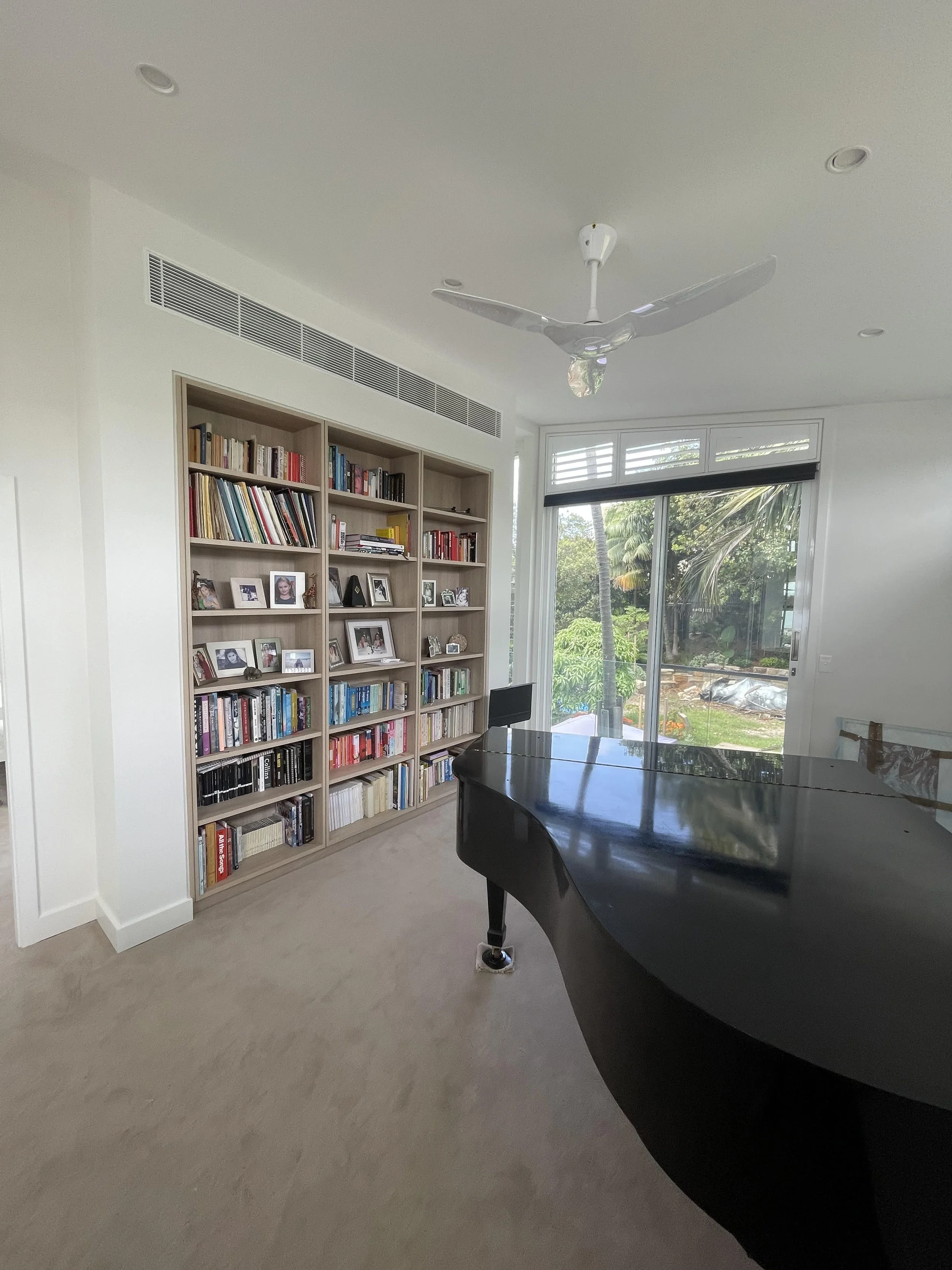 Living room with bookshelf, grand piano, ceiling fan, and large window showing outdoor greenery.
