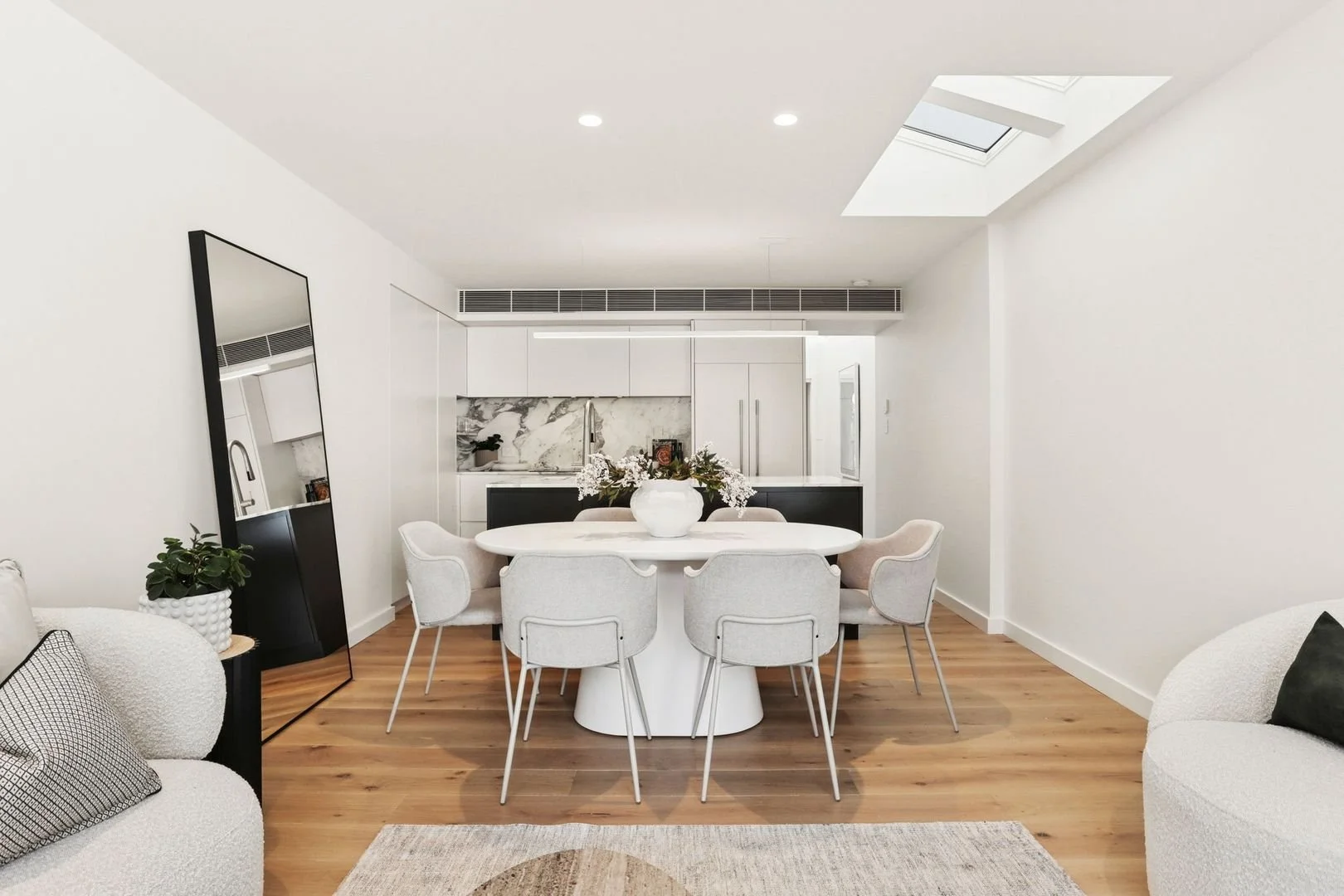 Modern, minimalistic living and dining area with white walls, wooden flooring, a rectangular mirror leaning against the wall, armless chairs around a white dining table, a coffee table, and a modern kitchen with white cabinets and marble backsplash in the background.