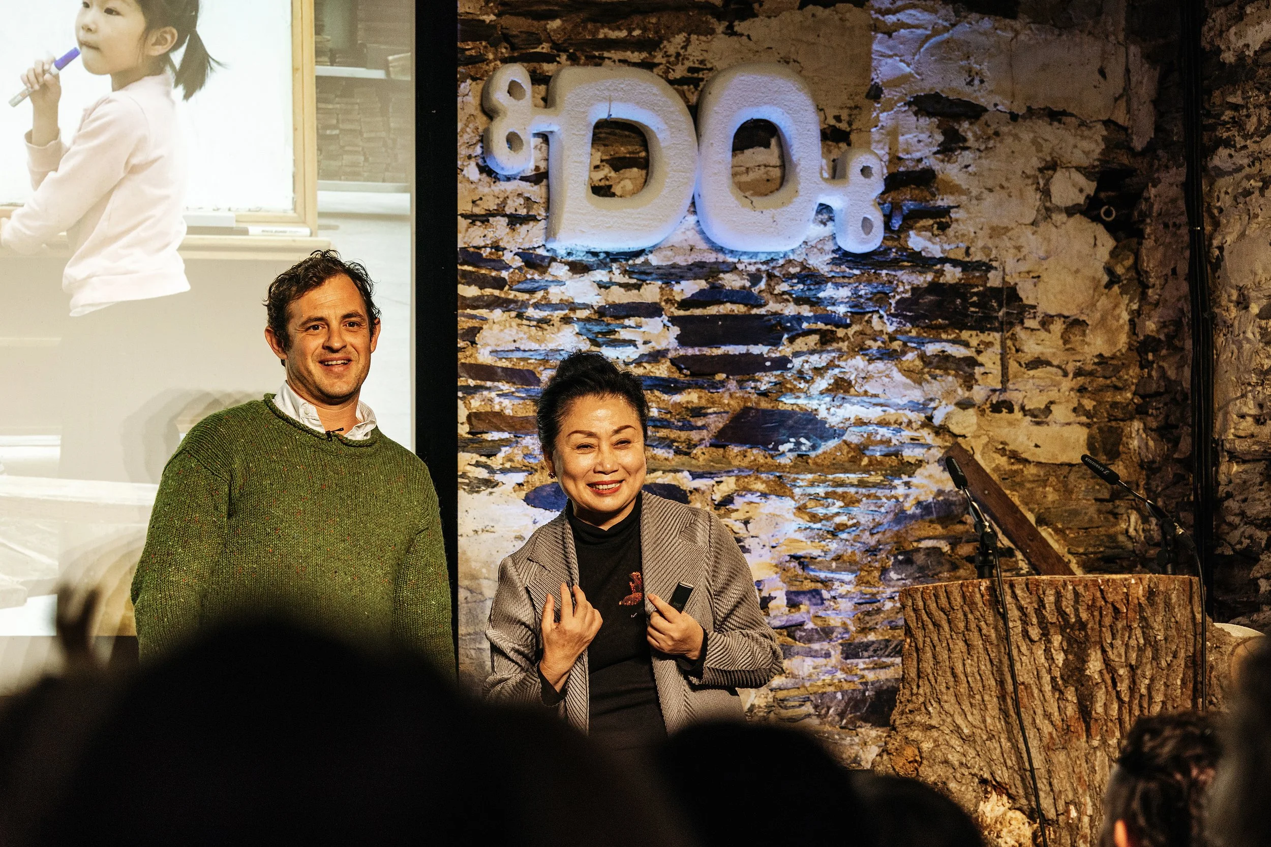 Two people standing on stage in front of a brick wall with a large sign that says 'DOK'. One is a man with curly hair wearing a green sweater, and the other is a woman with dark hair in a bun wearing a gray blazer, smiling.