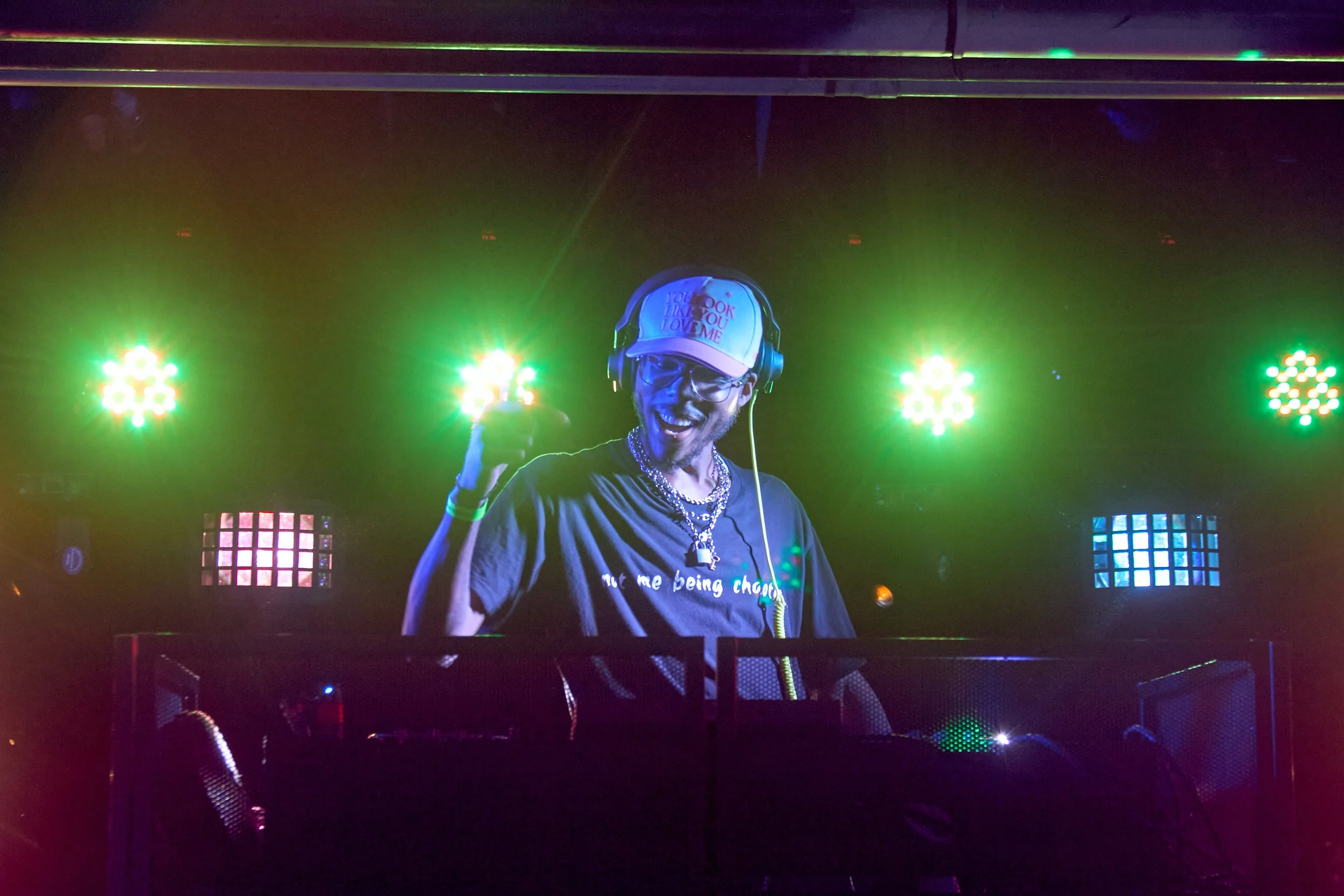 A DJ wearing glasses, a white cap that says "You Look Like You Love Me," and headphones, smiling while performing on stage with colorful lights in the background.