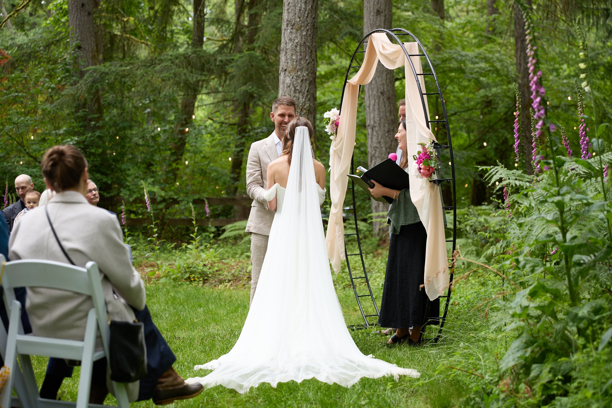 Outdoor wedding ceremony in a forested area with a bride and groom exchanging vows under a decorated arch. Guests seated nearby, greenery, and trees surrounding the scene.