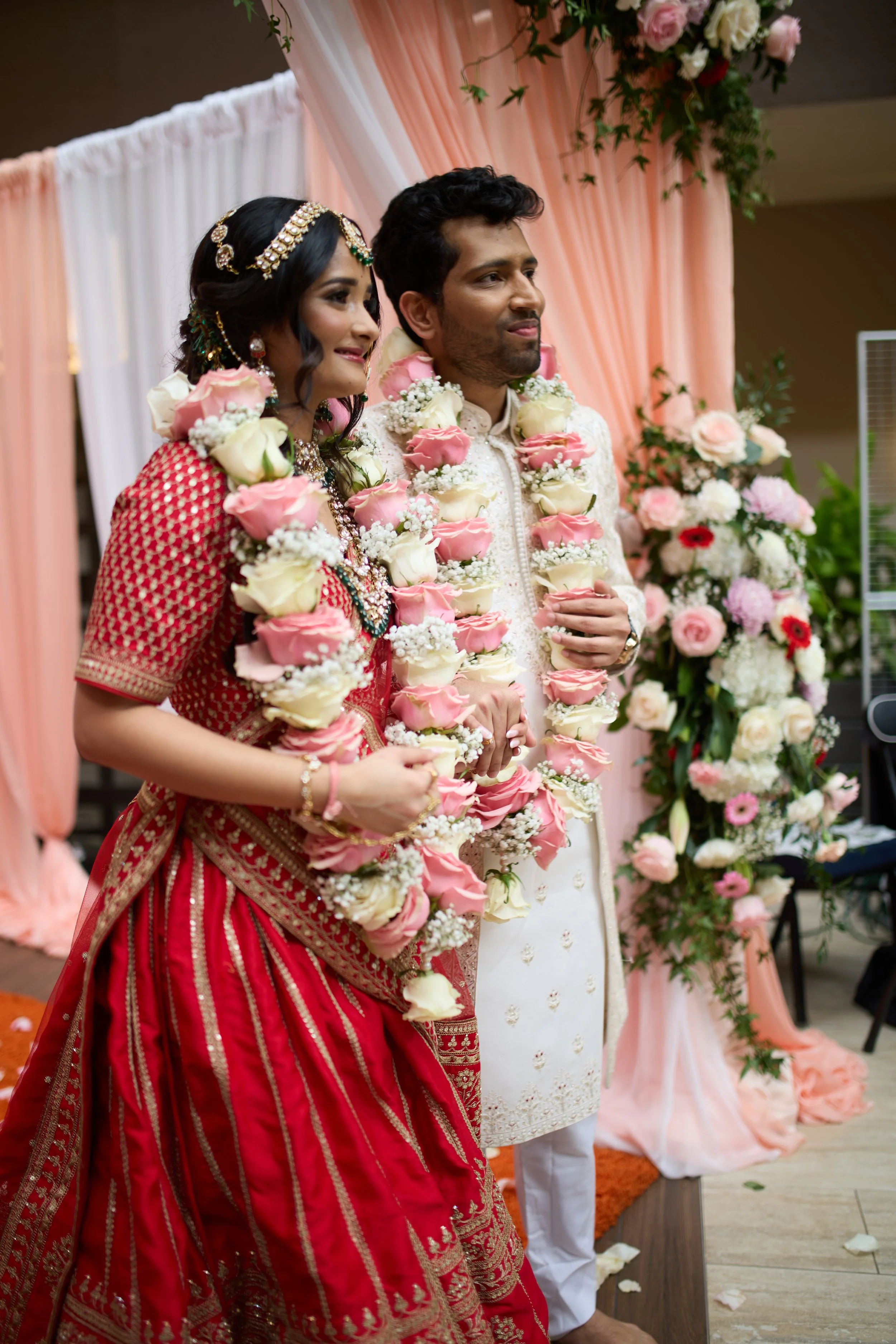 A couple dressed in traditional Indian wedding attire with garlands of pink and white roses around their necks, standing in front of a floral backdrop at a wedding ceremony.