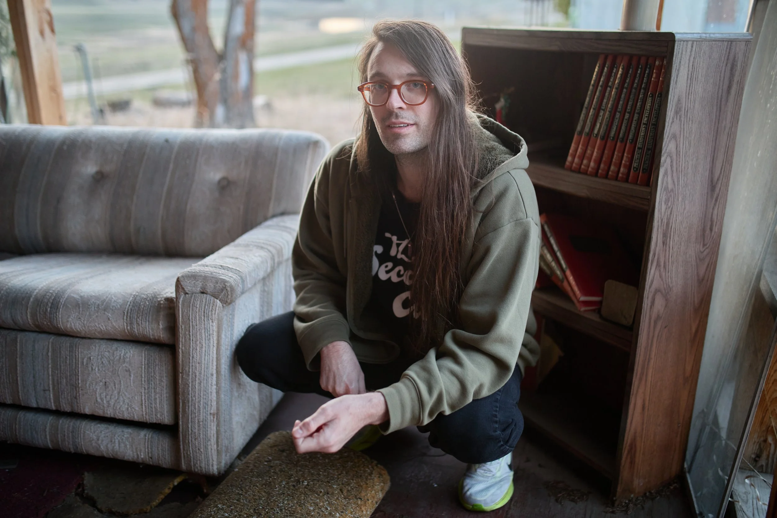 A person with long brown hair, glasses, and a green jacket is kneeling on the floor and arranging rocks in front of a vintage sofa in a cozy, rustic room with large windows.